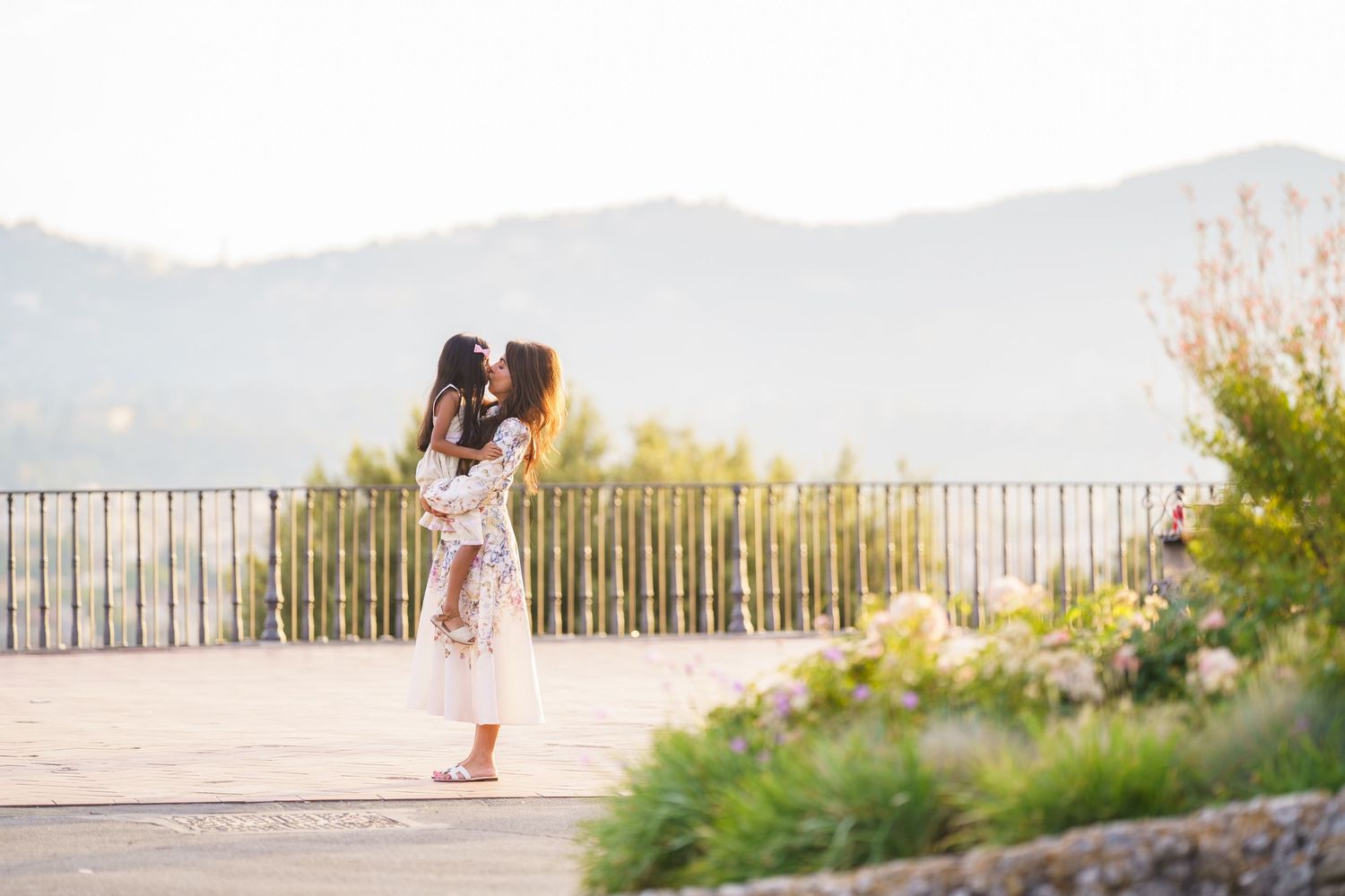 Mother kissing her child outdoors on a scenic terrace with mountain views and lush greenery.