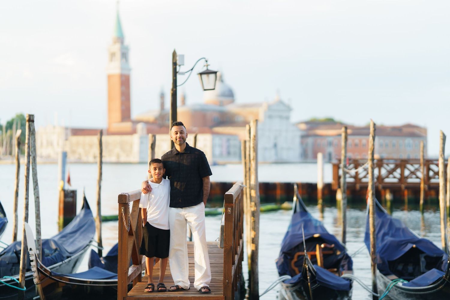 Father and son standing on a wooden dock in Venice, Italy, with gondolas and San Giorgio Maggiore in the background.