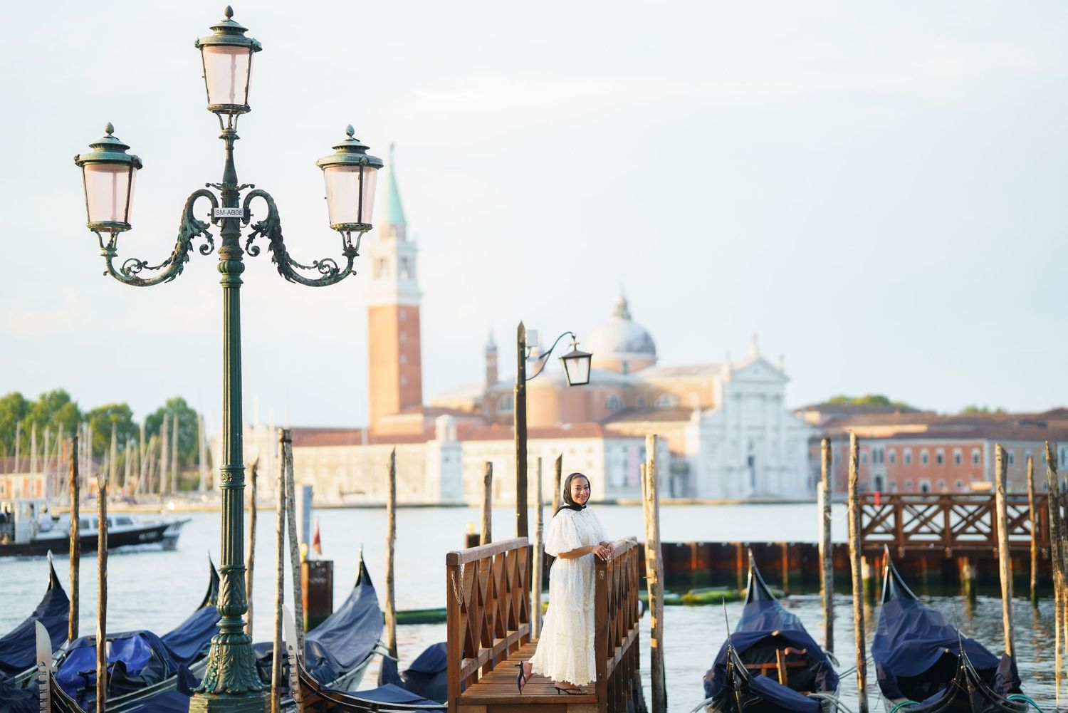 Woman in white dress standing on Venice gondola dock with San Giorgio Maggiore church and lagoon in background.