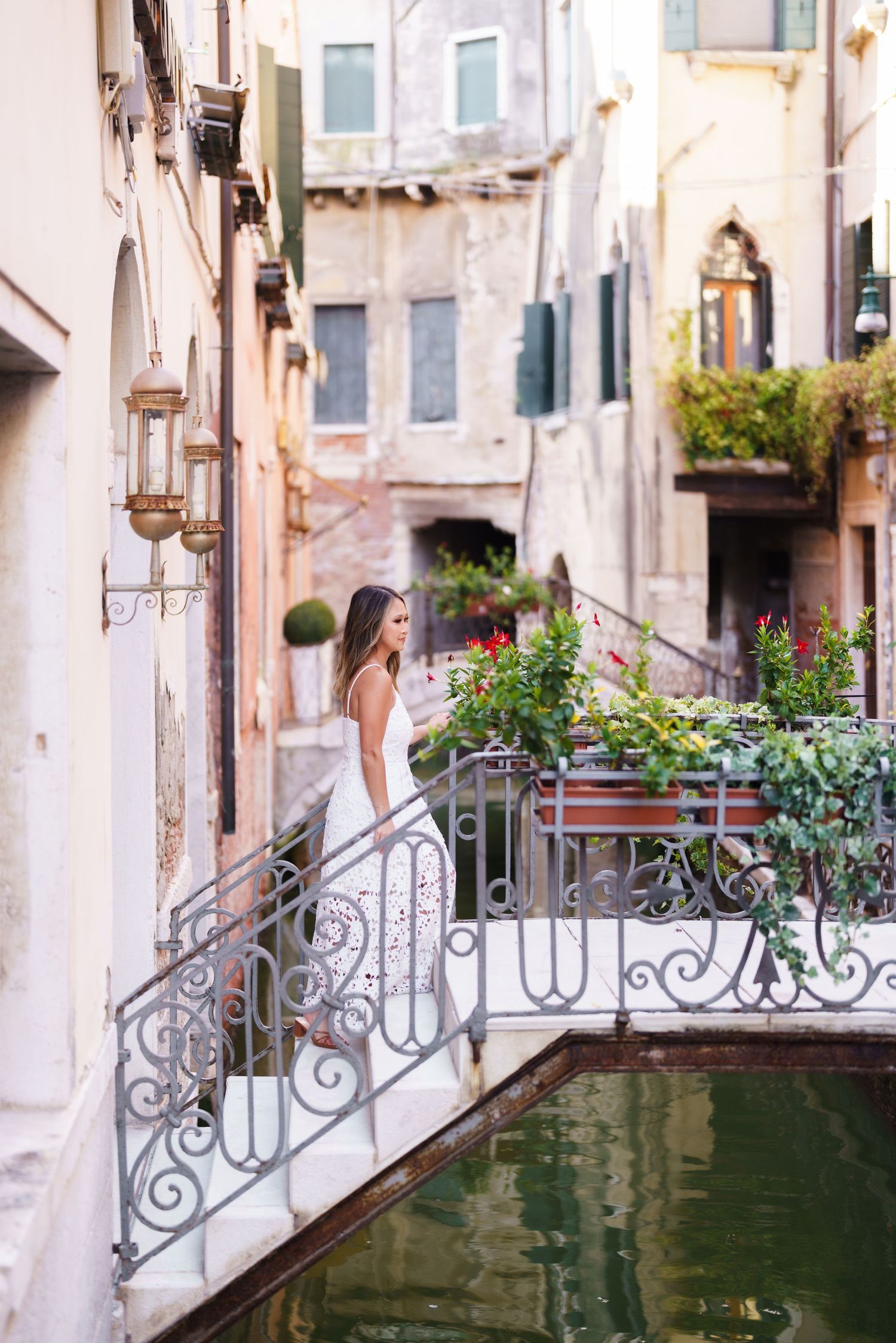 Woman in white dress standing on ornate bridge over Venice canal with colorful flowers and historic buildings.