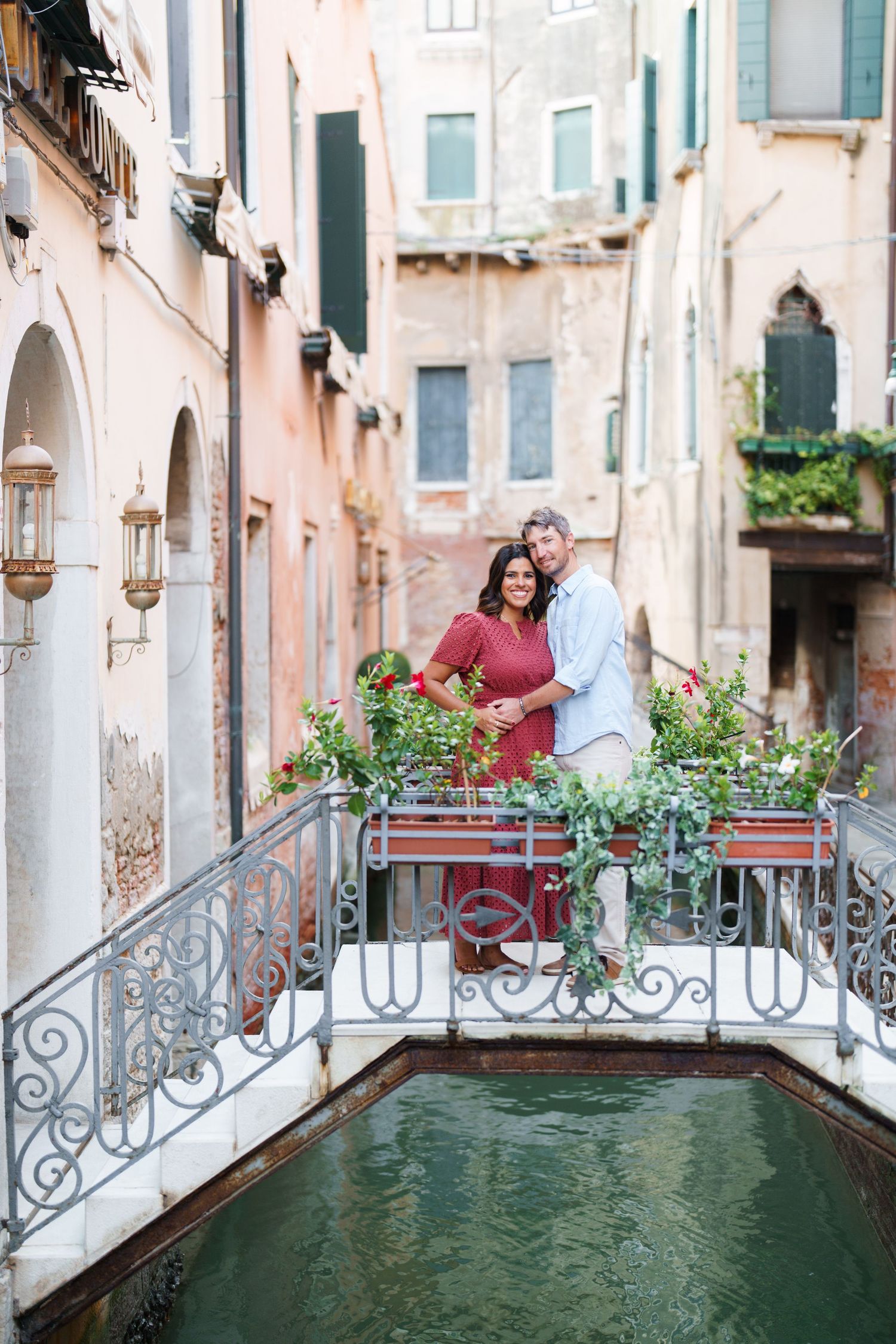 Couple embracing on ornate bridge over Venice canal surrounded by colorful flowers and historic buildings.