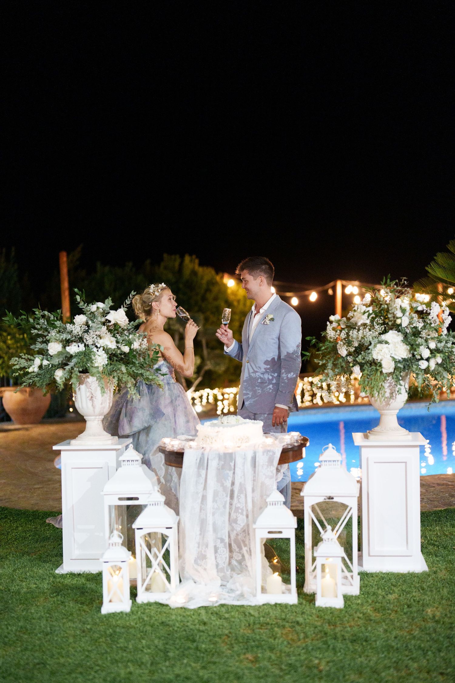 Bride and groom toasting with champagne at elegant outdoor night wedding reception with white lanterns and floral arrangements.