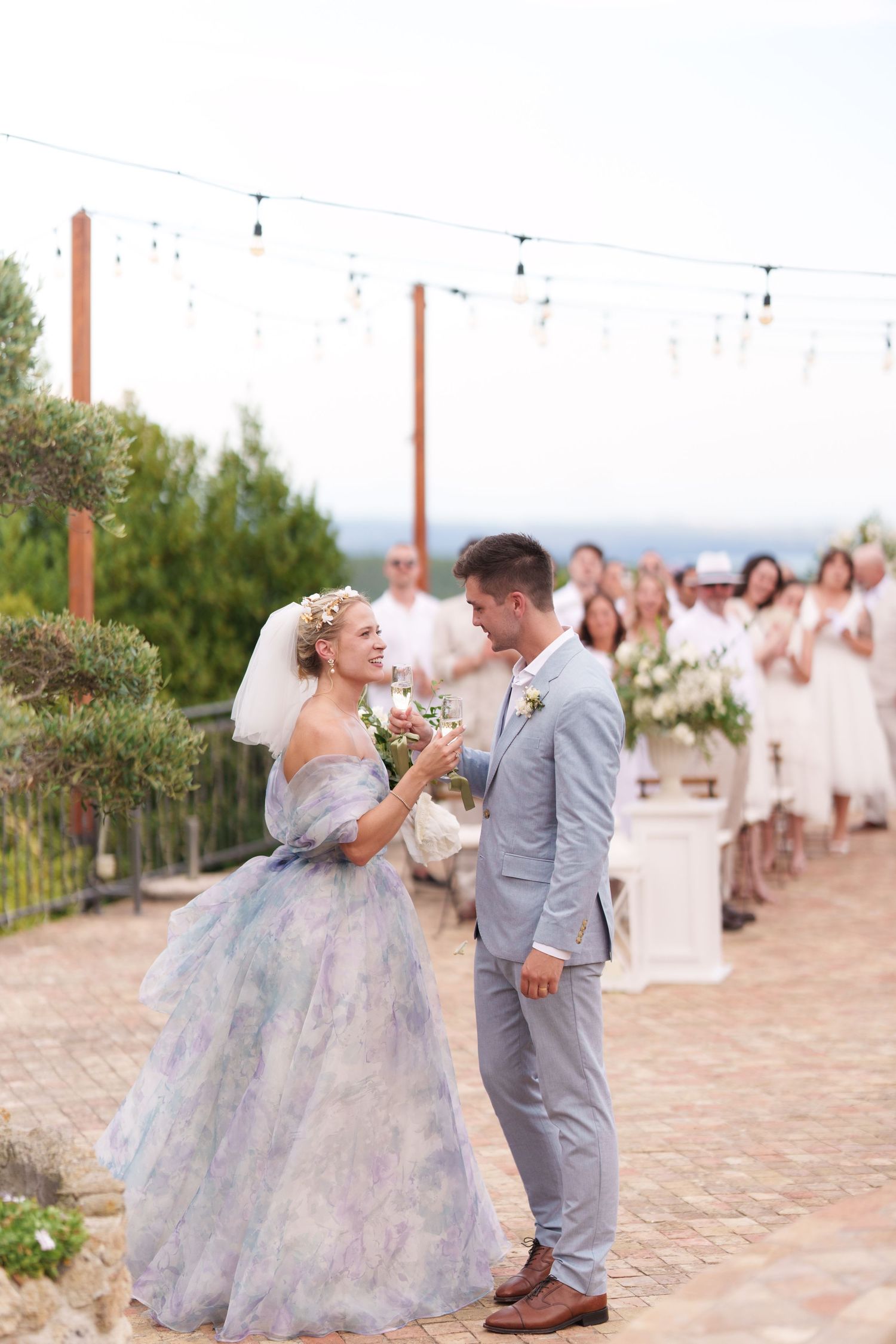 Bride in floral gown and groom in gray suit share a romantic moment at an outdoor wedding ceremony.