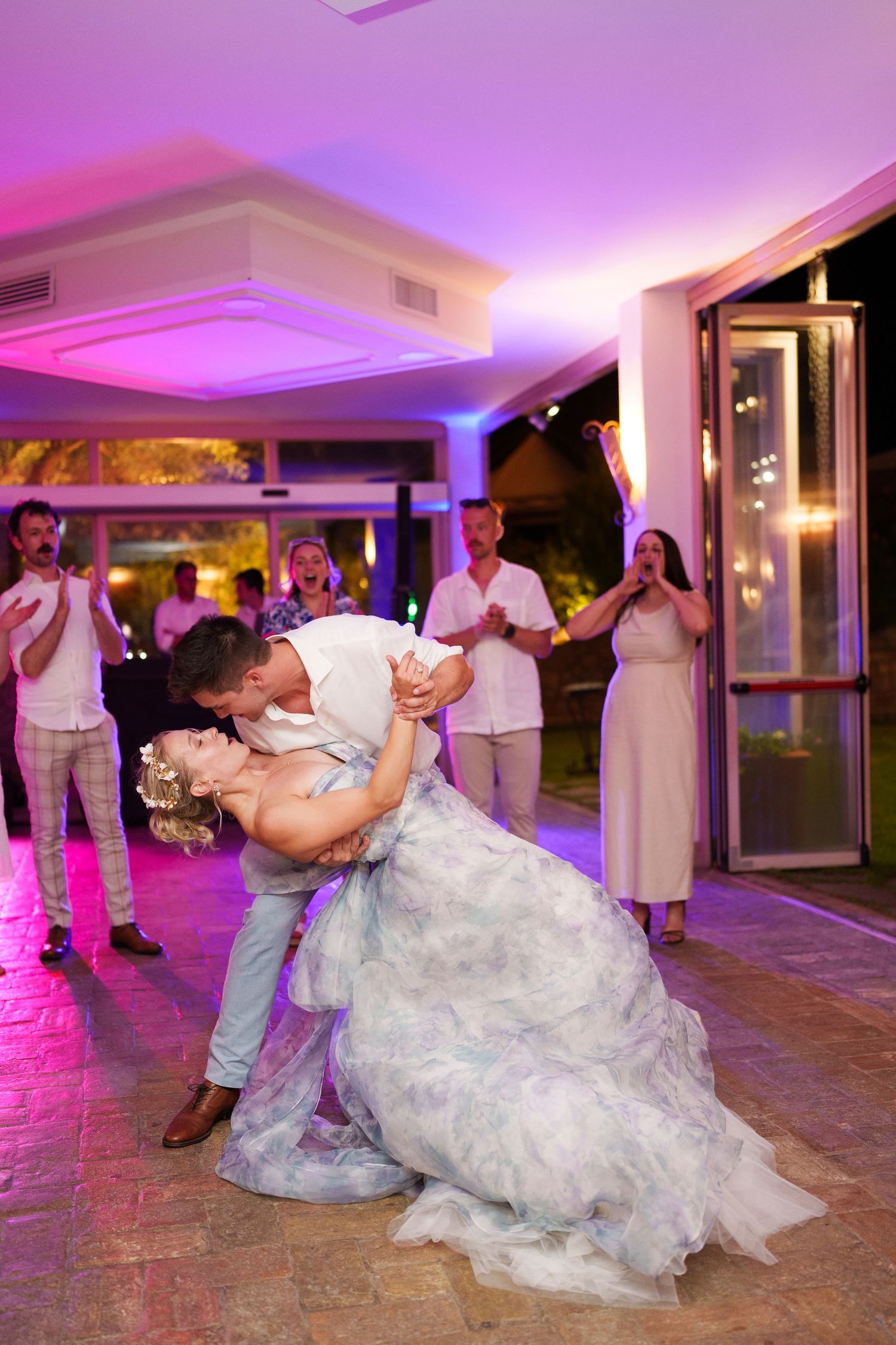 Bride and groom share a romantic dip kiss on the dance floor while wedding guests cheer and applaud behind them.