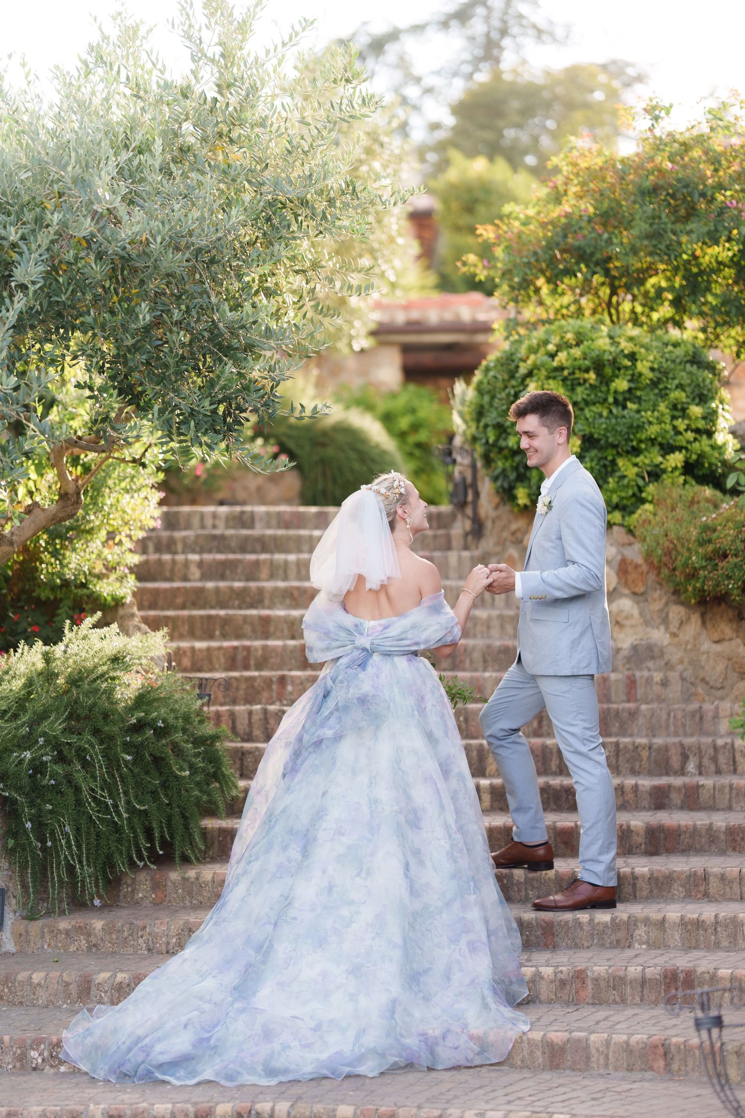 Bride in blue floral gown and groom in gray suit hold hands on garden steps at outdoor wedding.