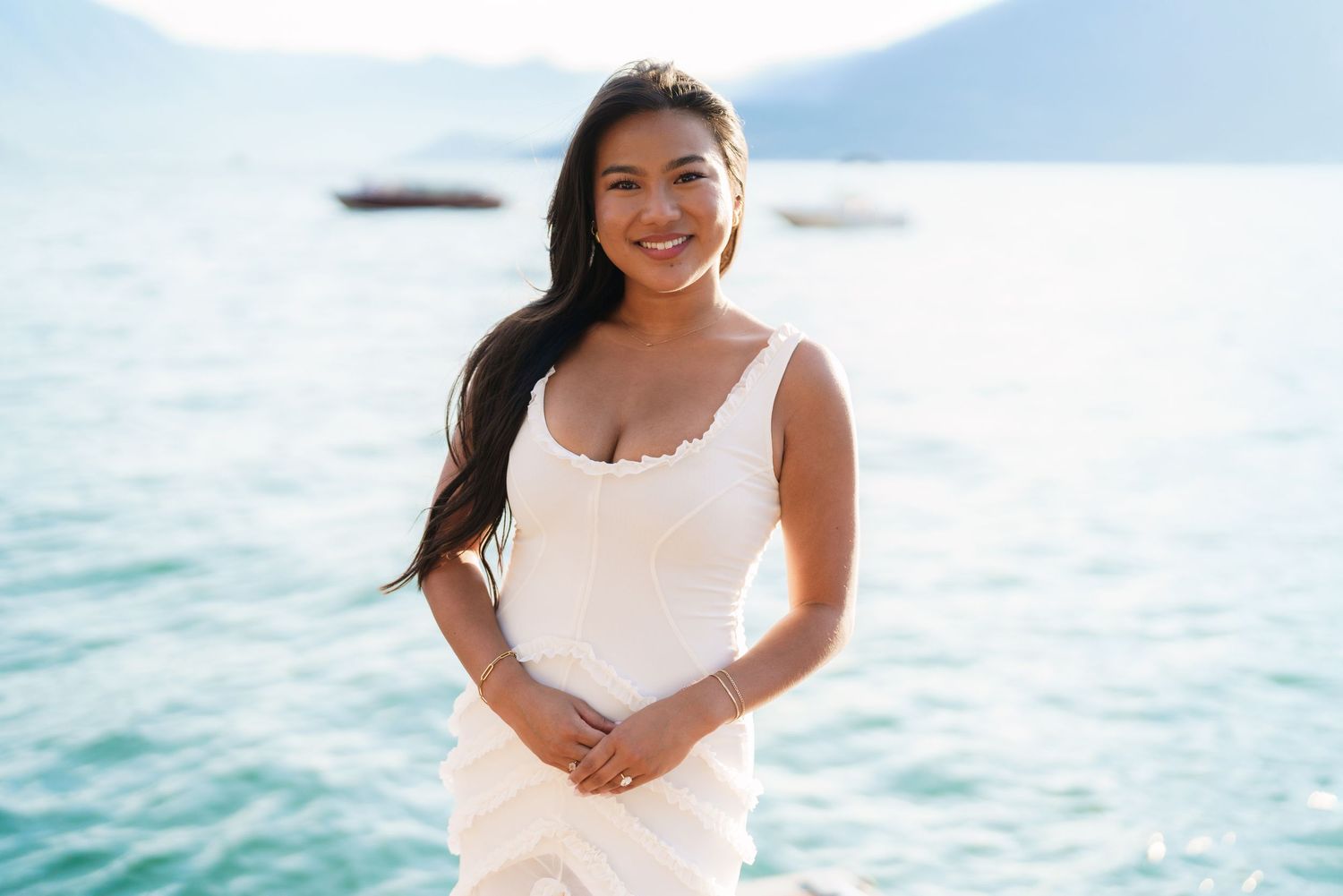 A smiling young woman in a white dress poses by a turquoise lake with mountains in the background.