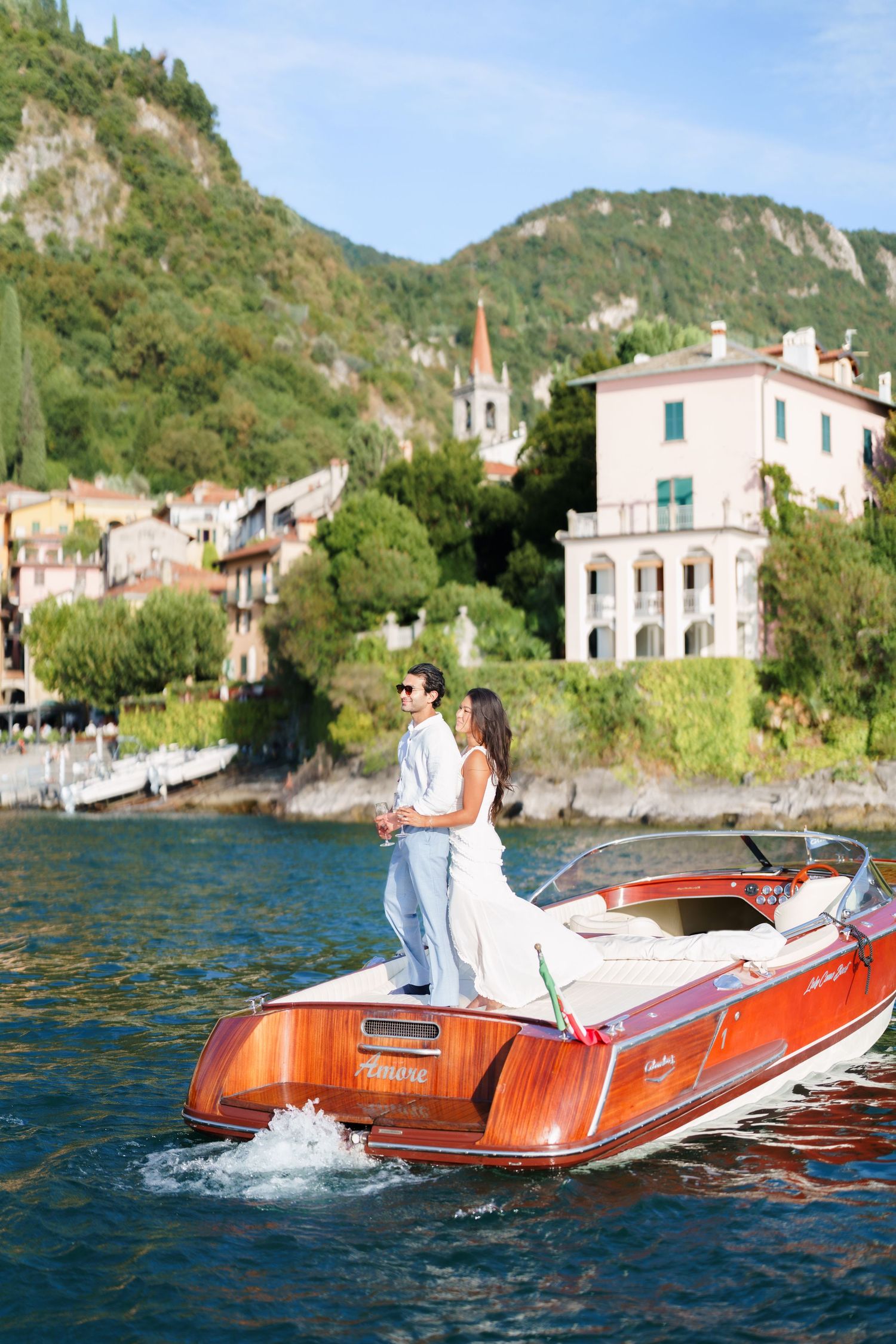 Couple standing on a classic wooden Riva speedboat on Lake Como, Italy, with a pink villa and green hills behind them.