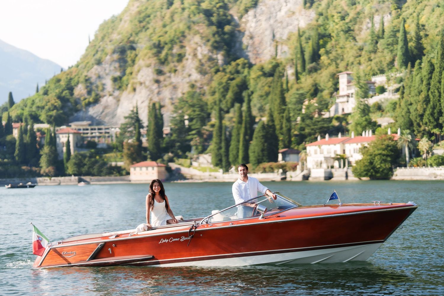 Couple riding a classic red wooden speedboat on Lake Como, Italy, with lush hillside villas in the background.