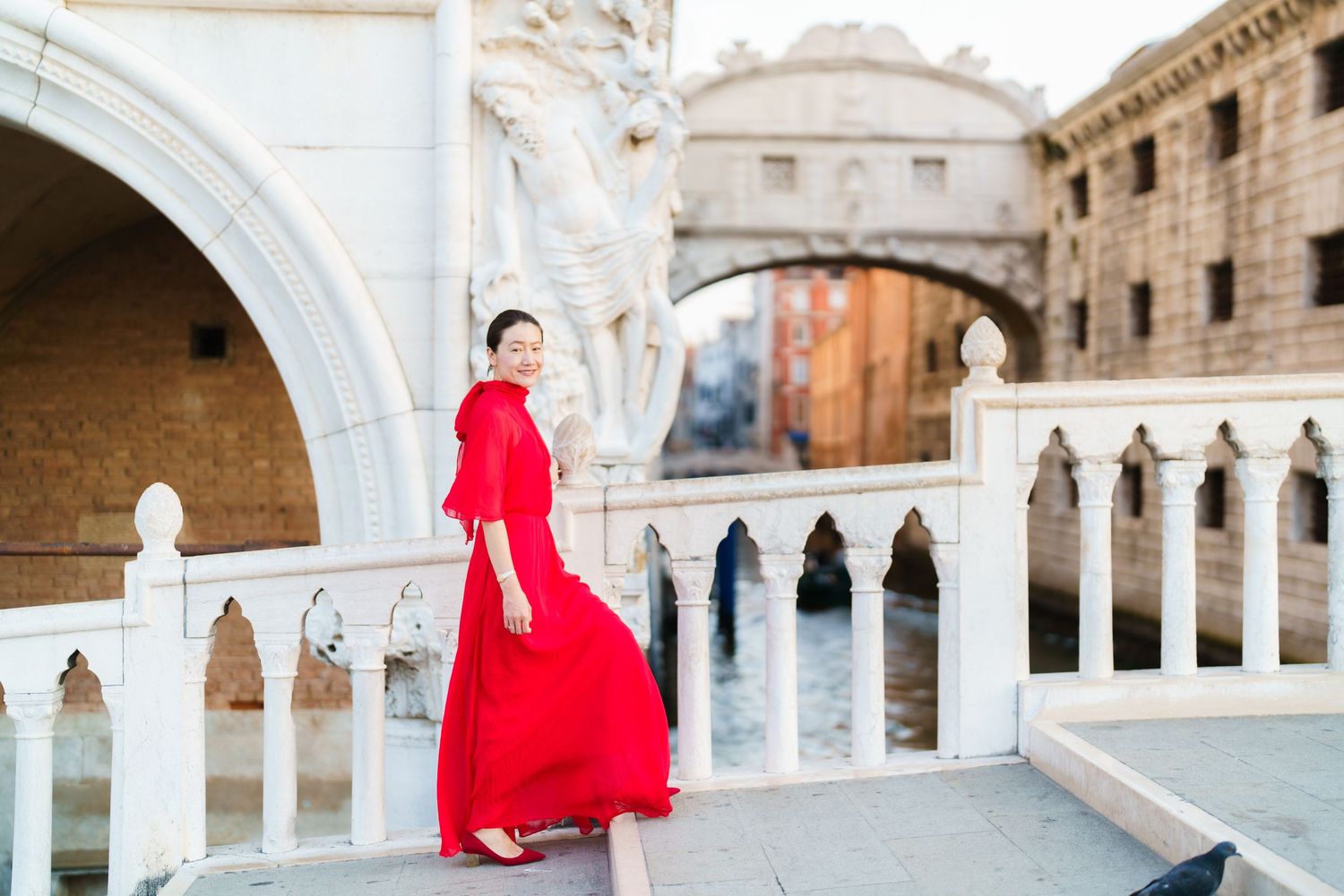 Woman in red flowing dress posing on a Venice bridge with the Bridge of Sighs and canal in background.