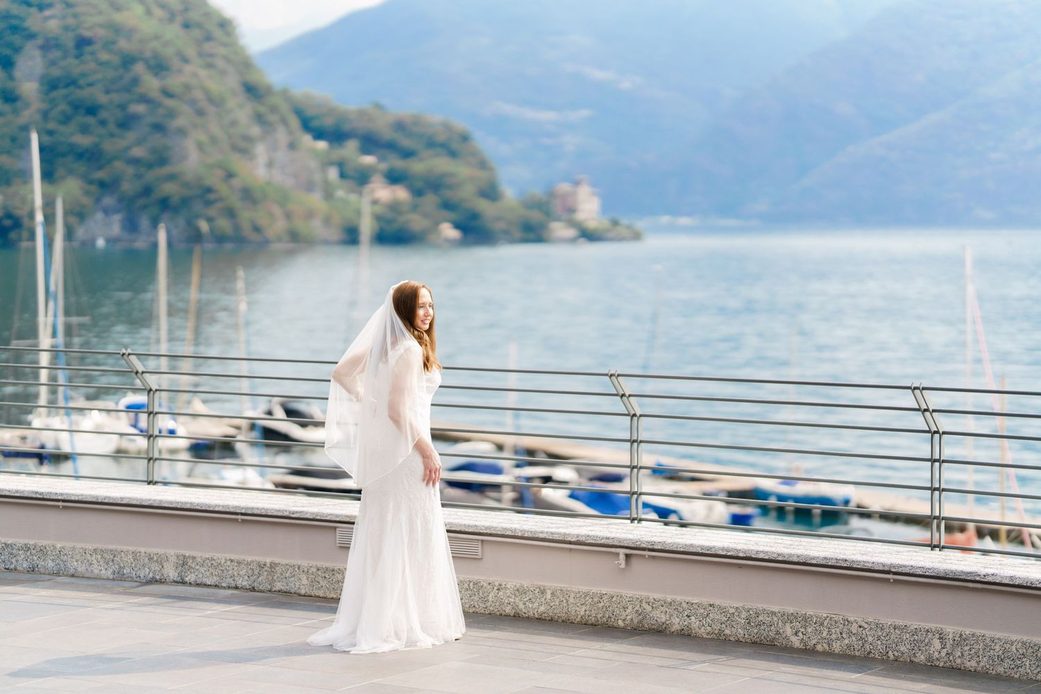 Bride in white gown and veil stands by lakeside railing with boats and mountains in background.