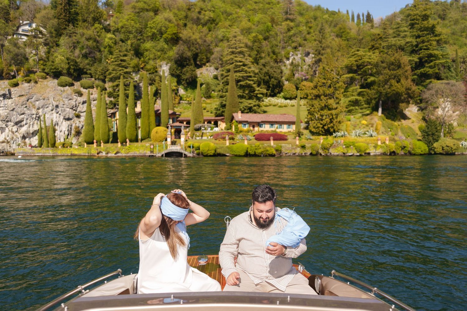Couple on a classic wooden boat on a scenic lake with lush green hillside villa in the background.