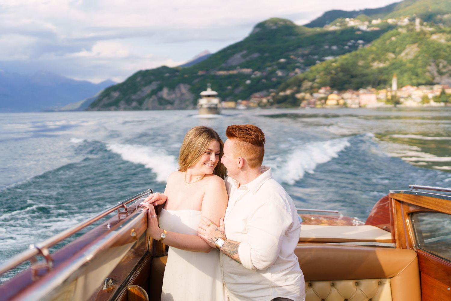 Couple embracing on a wooden boat on Lake Como, Italy, with mountain scenery and sparkling water wake behind them.
