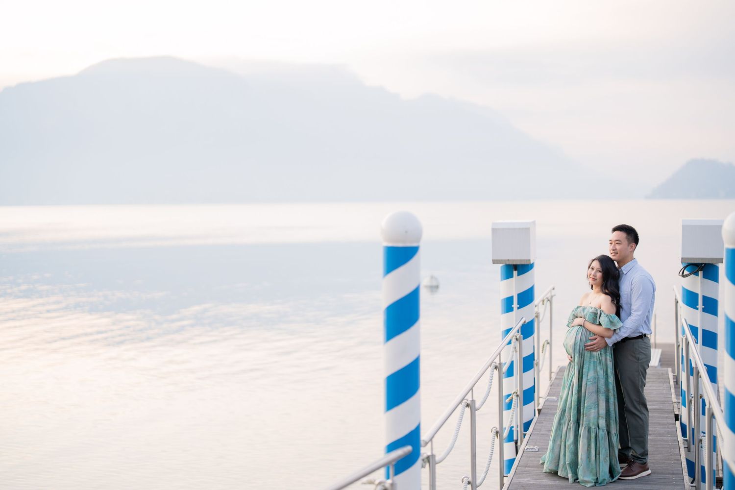 Couple embracing on a pier with blue and white striped poles, serene lake and misty mountains in the background.