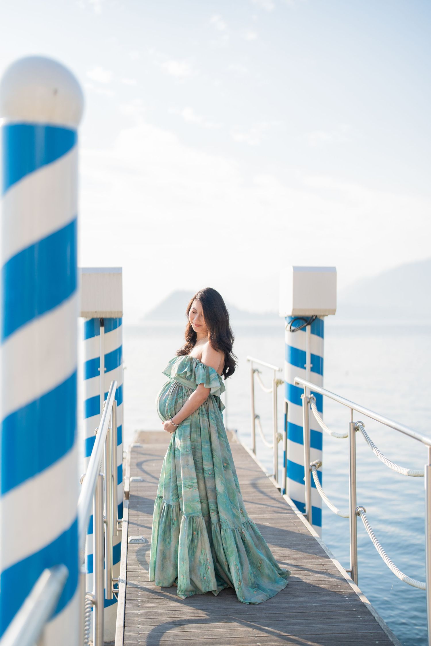 Pregnant woman in teal floral maxi dress standing on a pier with blue striped poles overlooking calm ocean waters.