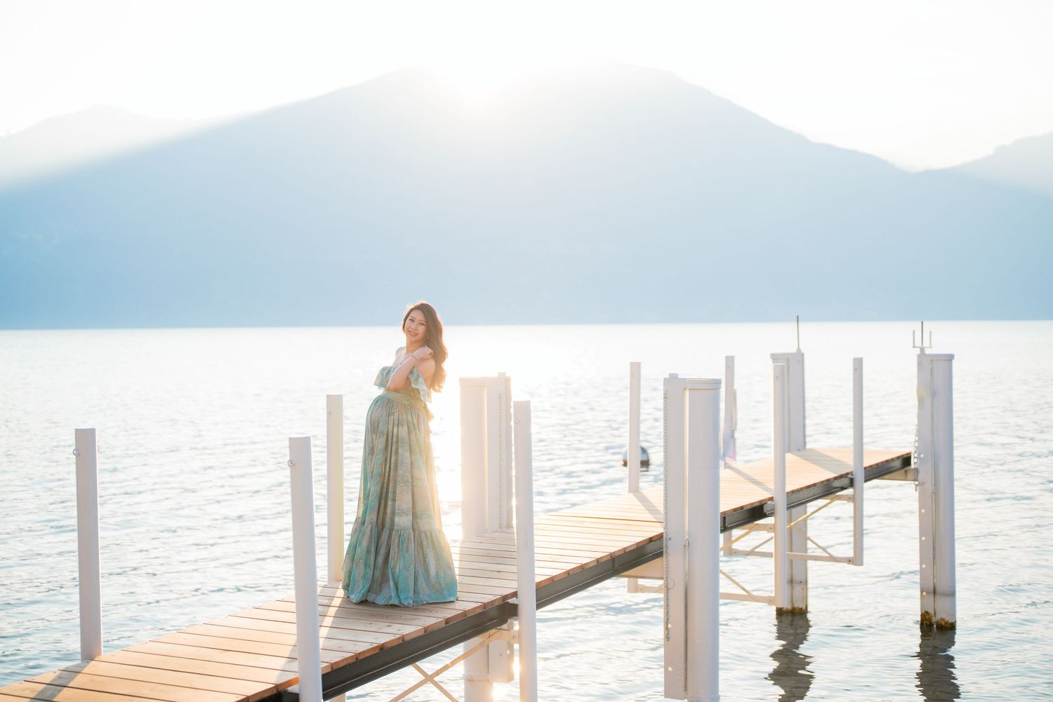 Woman in teal gown stands on a sunlit wooden dock overlooking a serene mountain lake.