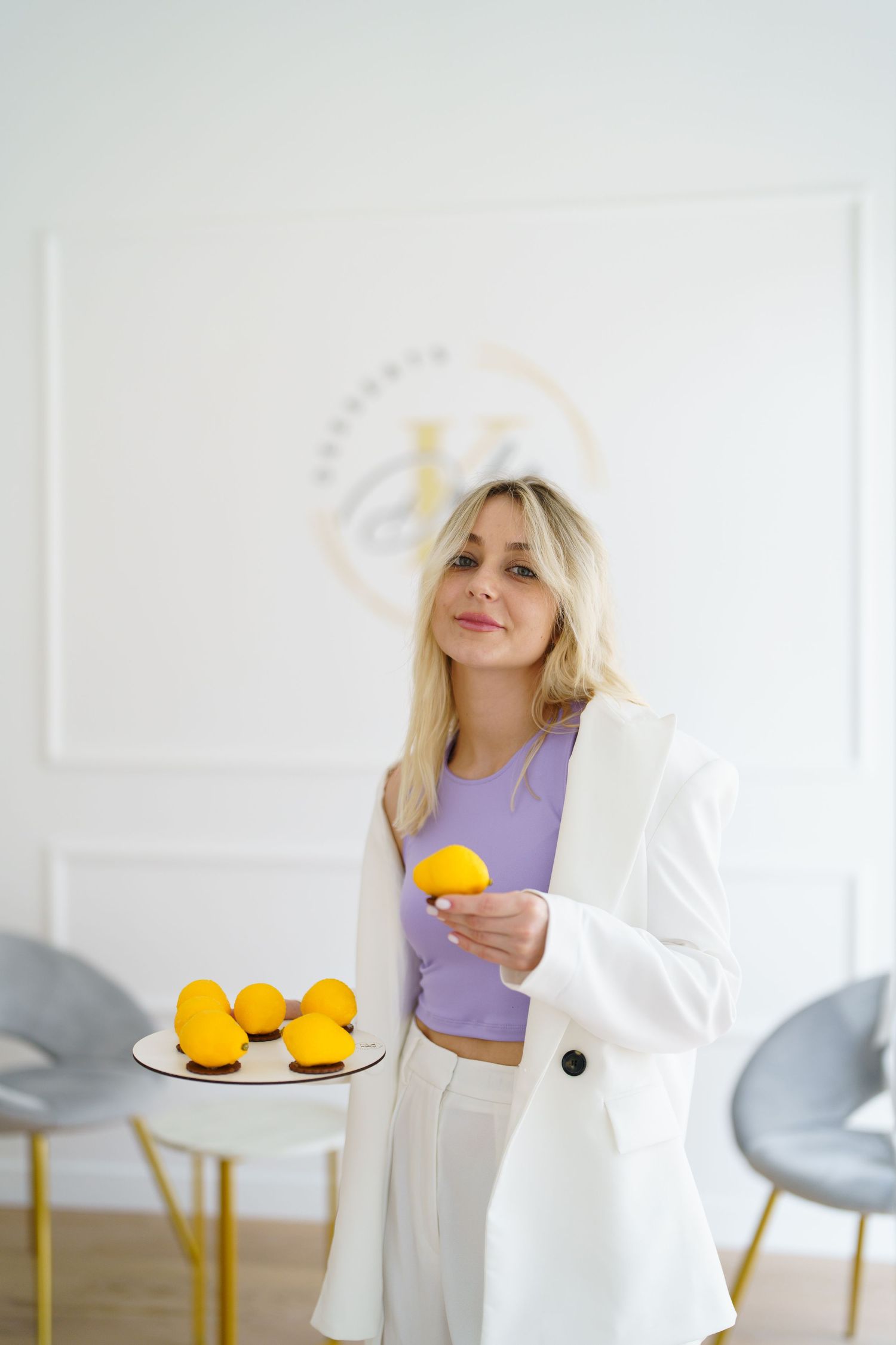 Blonde woman in white blazer and purple top holding a lemon, with a plate of lemons on a table in a bright modern room.