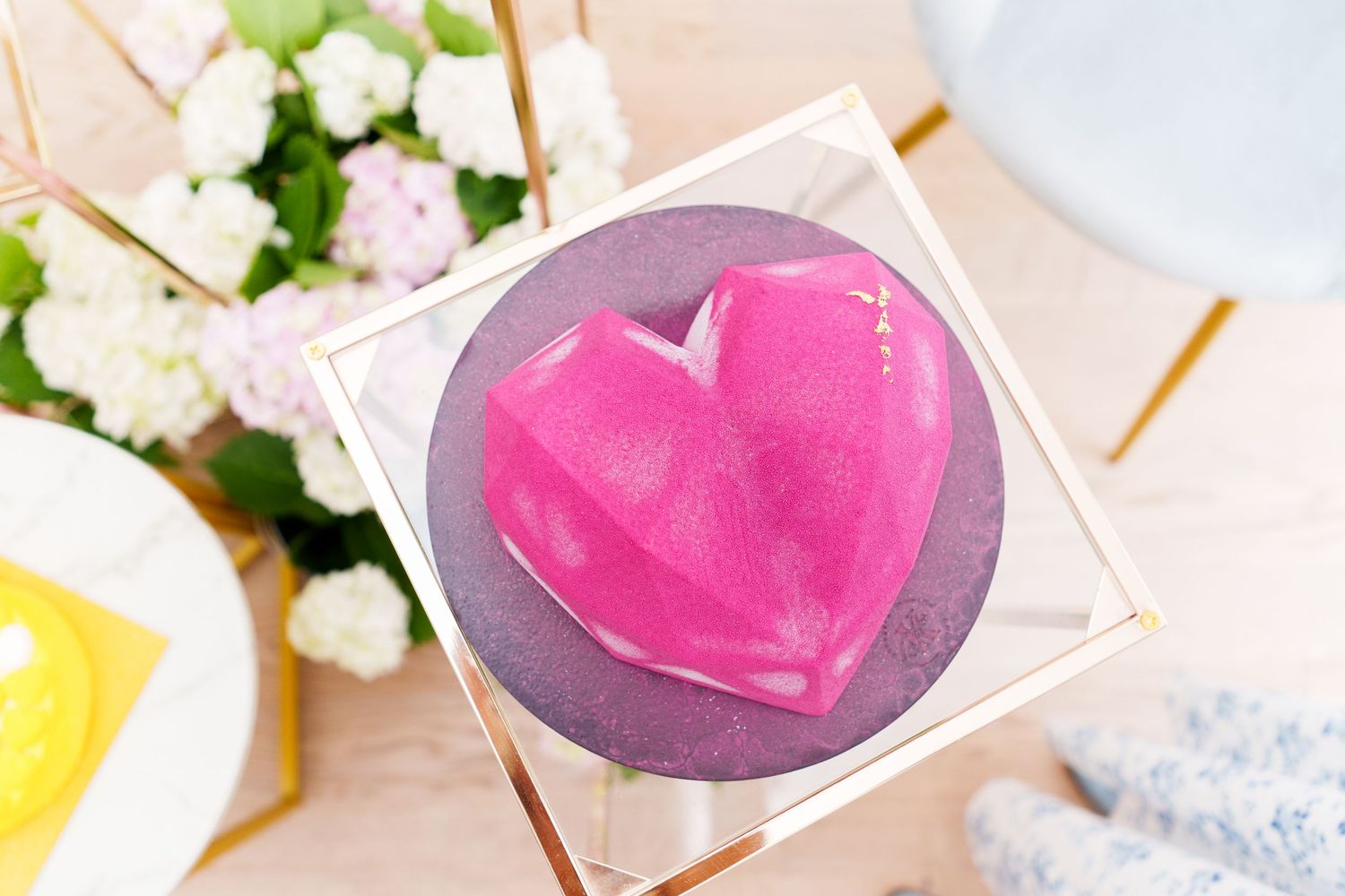 Pink geometric heart-shaped soap on a round purple base displayed in a gold acrylic box with flowers in background.