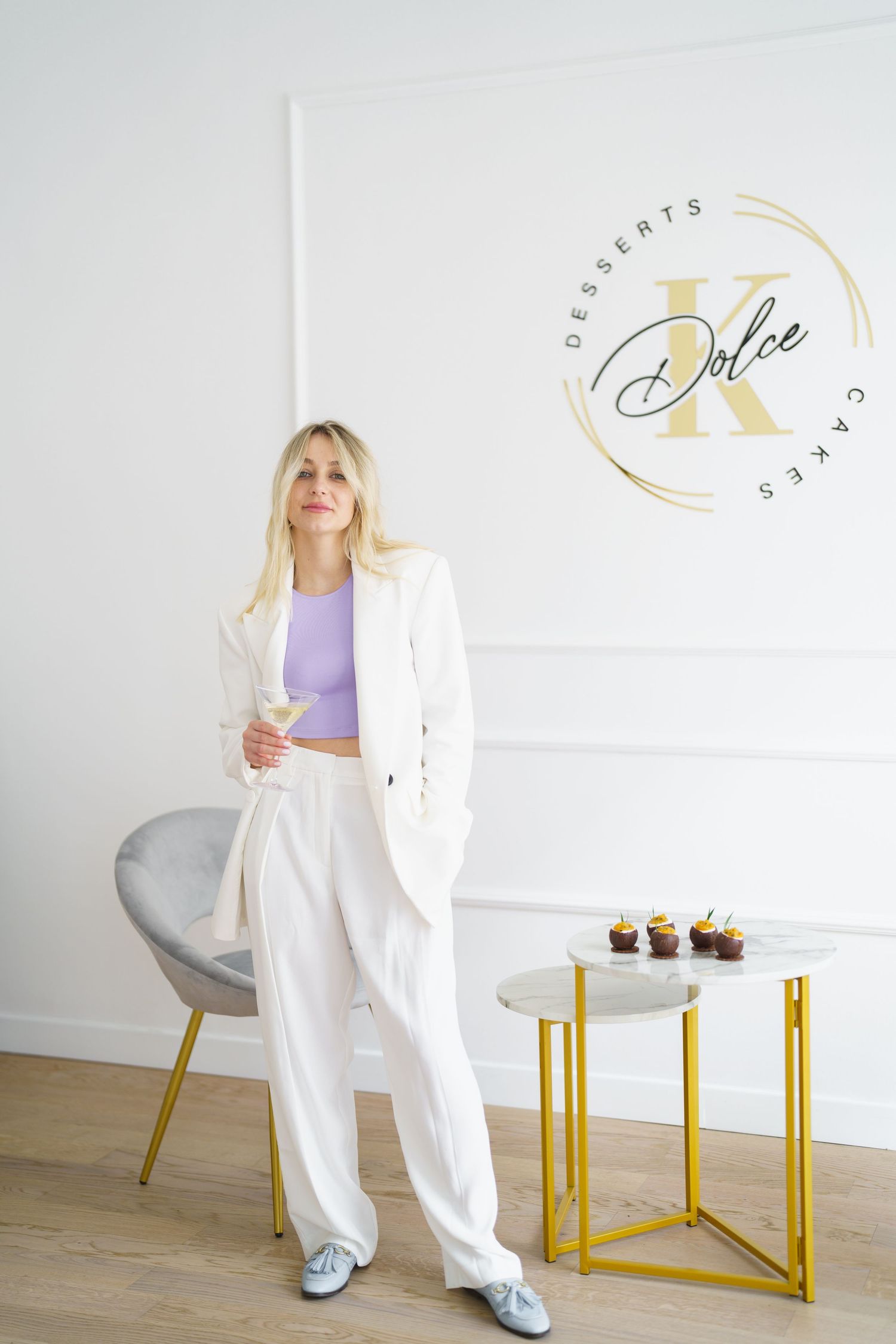 Blonde woman in white suit holding a glass near Dolce K Desserts Cakes logo with cupcakes on gold table.