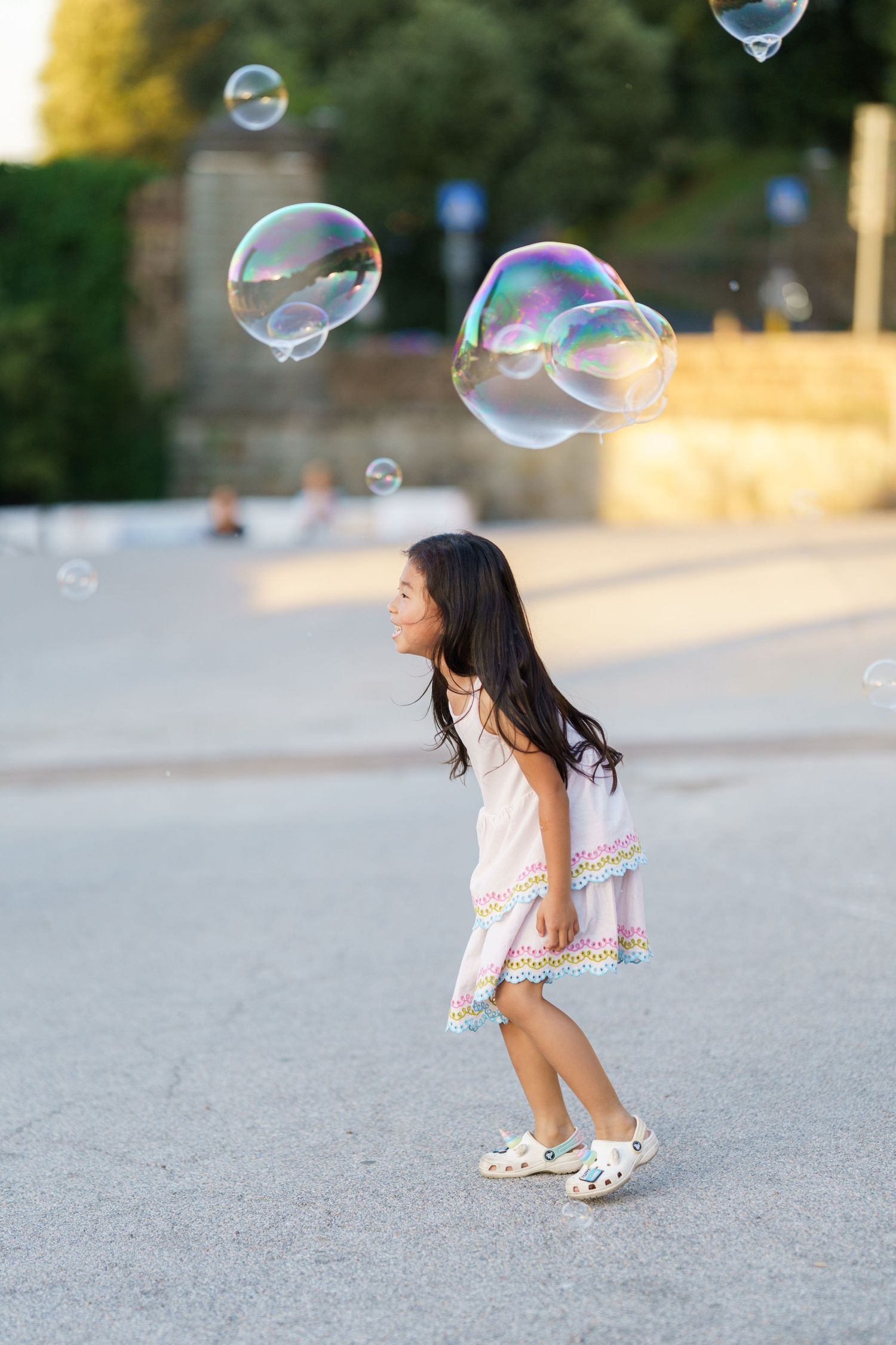 Young Asian girl in pink dress chasing large colorful soap bubbles outdoors in a sunny plaza.