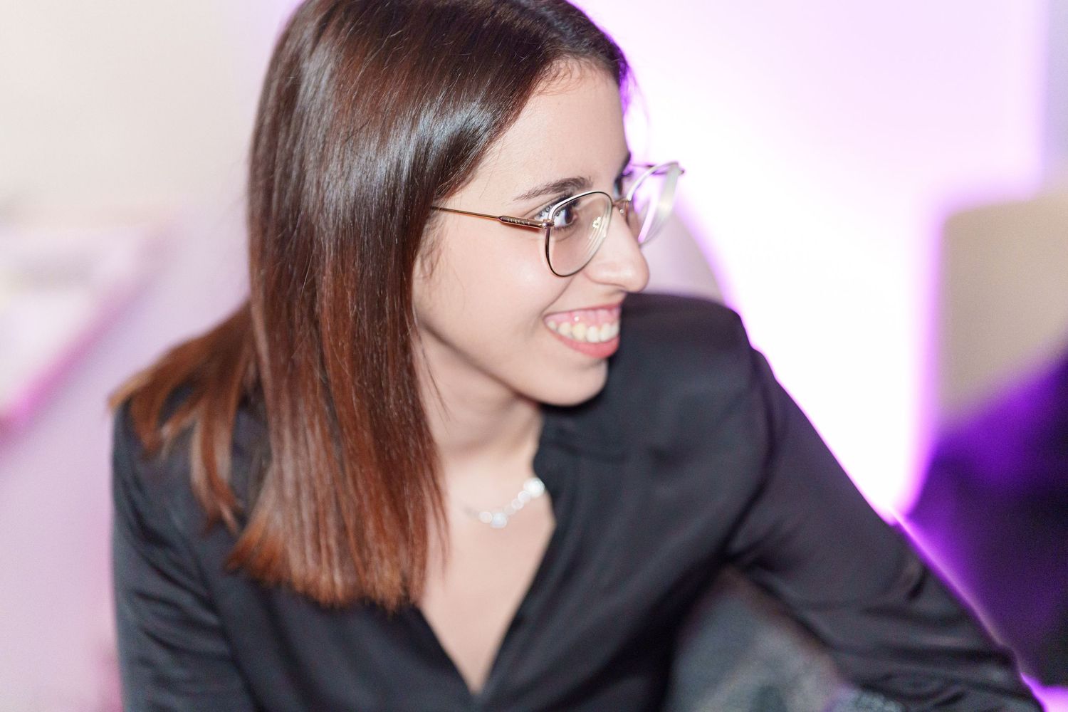 Young woman with glasses and dark hair smiling, wearing a black shirt in a softly lit setting.