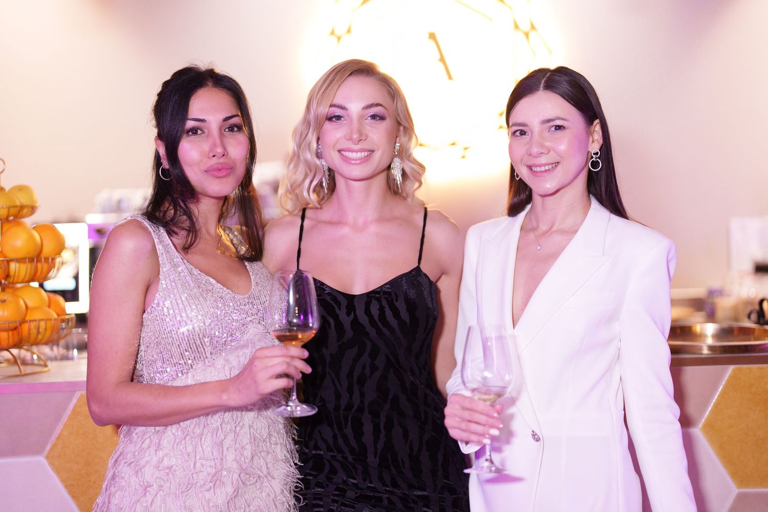 Three elegant women in formal attire holding champagne glasses pose together at a glamorous event.
