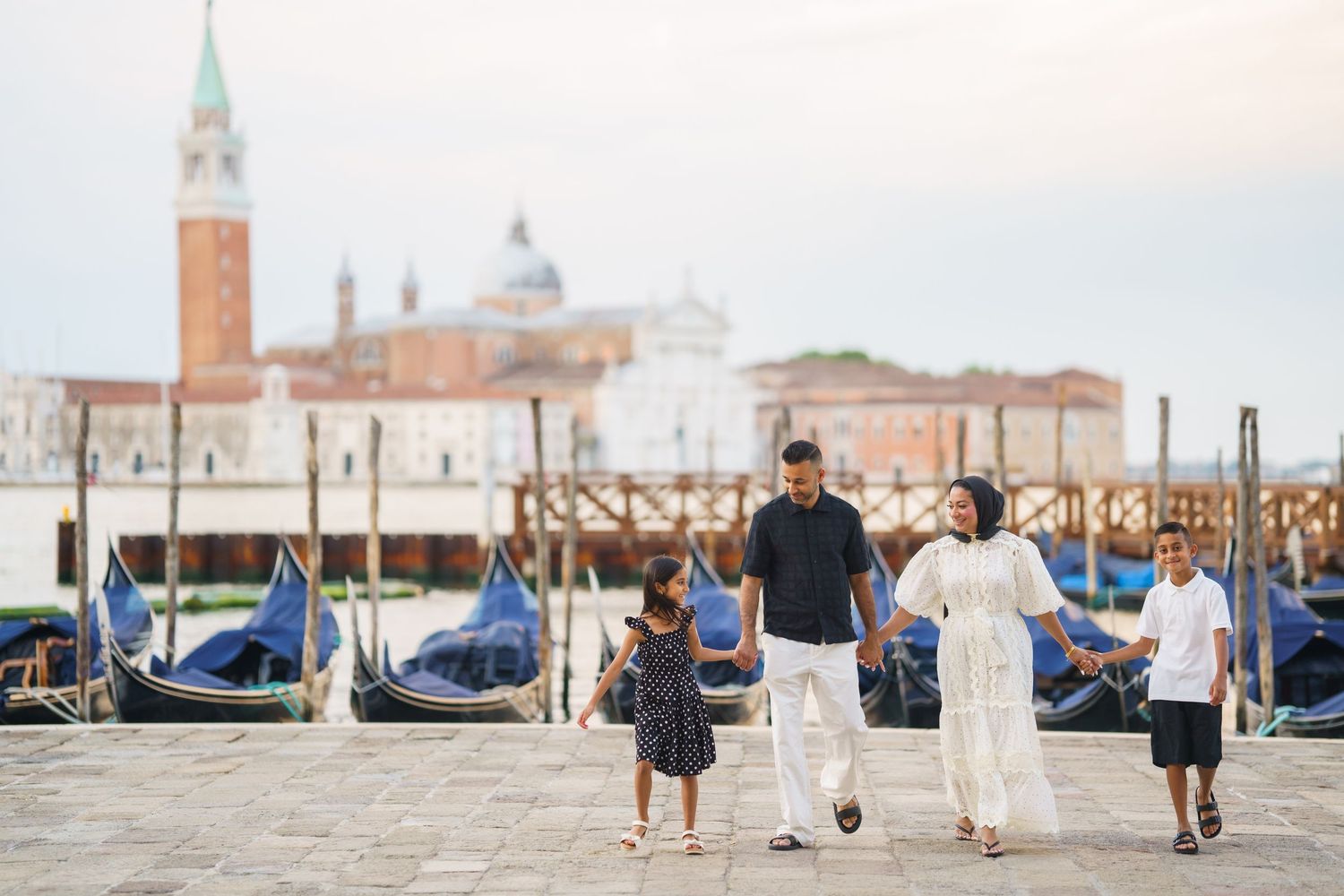Family of four walking along Venice waterfront with gondolas and San Giorgio Maggiore island in background.
