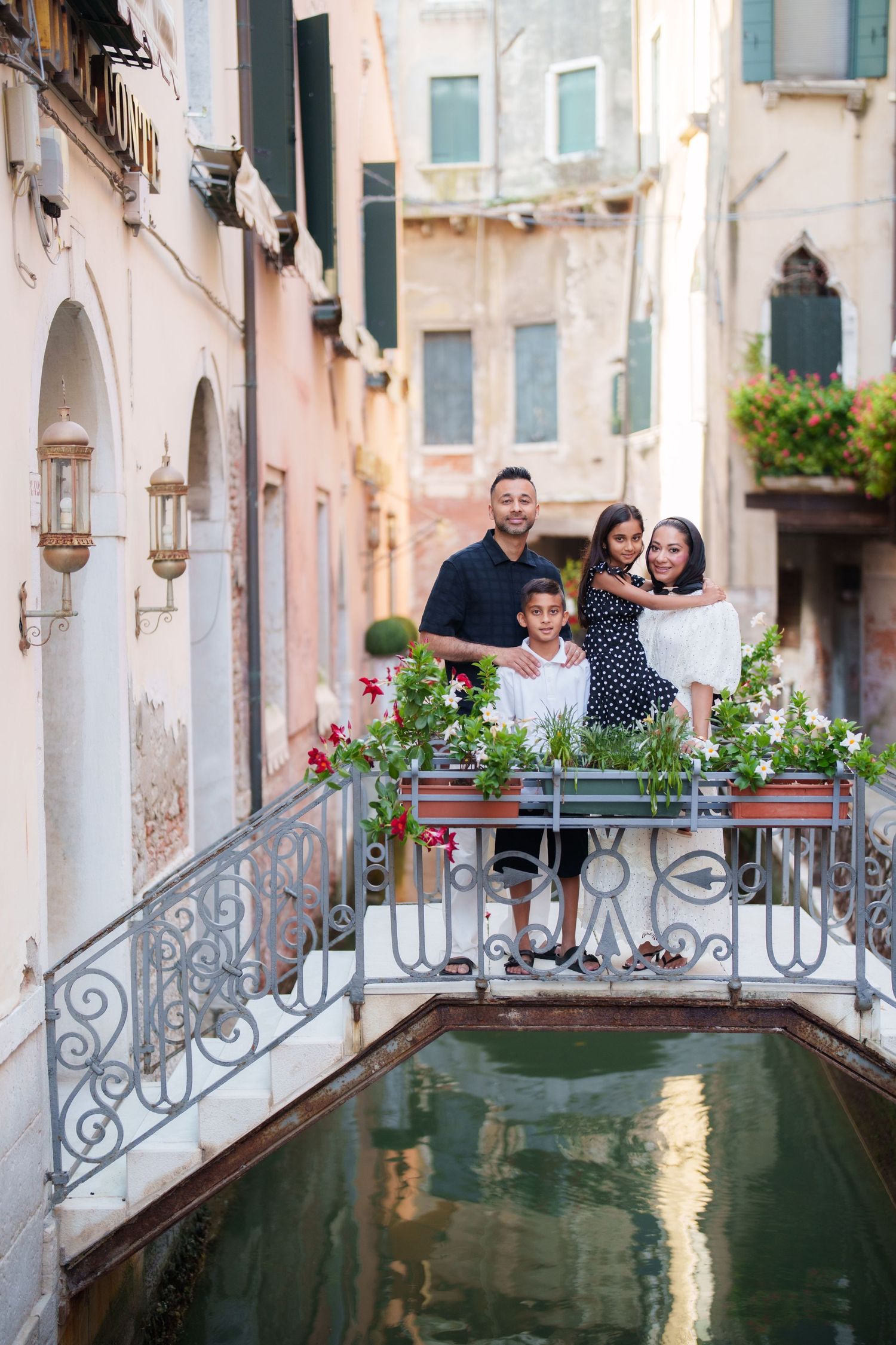 Family of three posing on a charming Venetian bridge over a canal, surrounded by colorful flowers and historic buildings.