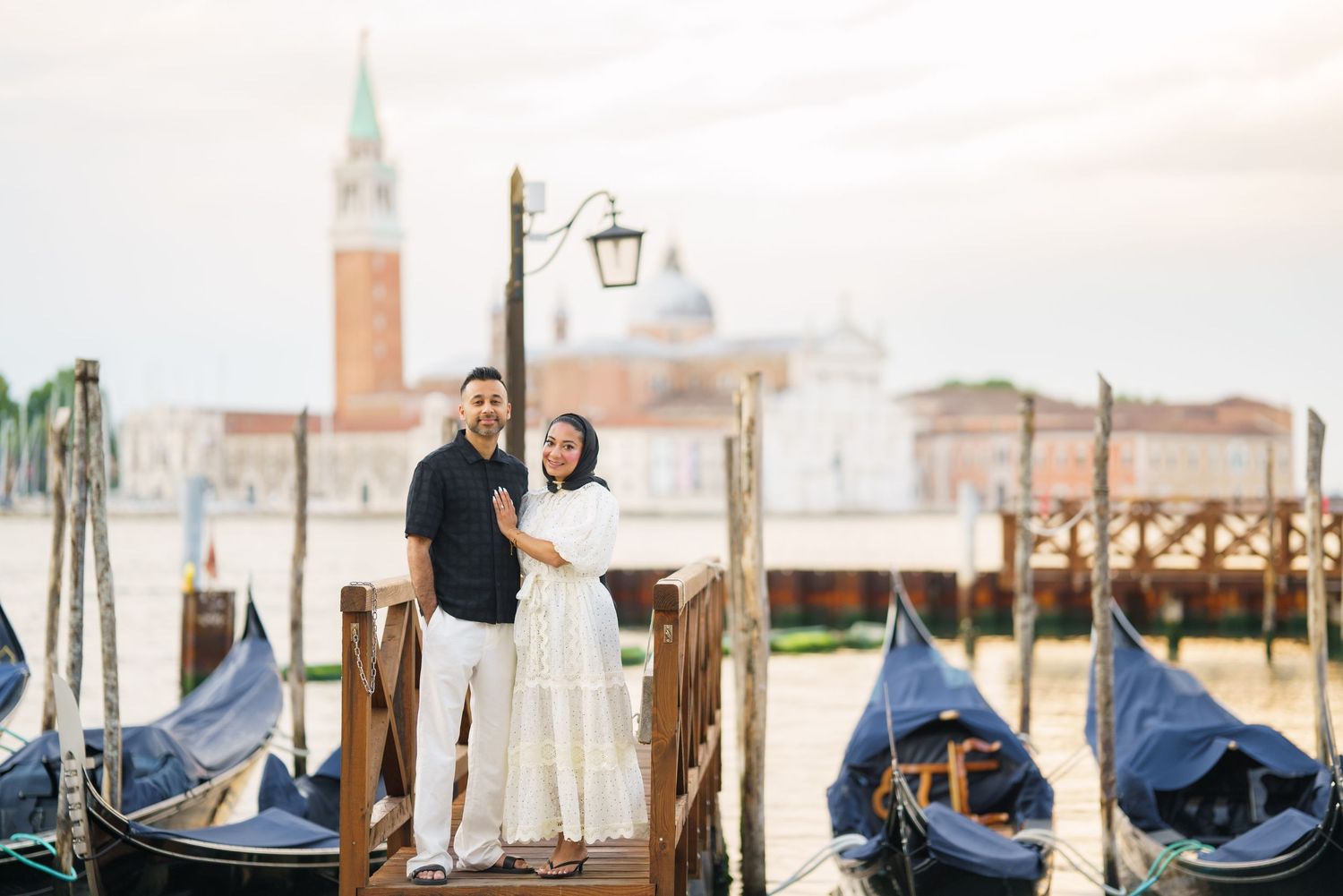 Couple posing on a wooden dock with gondolas and San Giorgio Maggiore church in Venice, Italy.