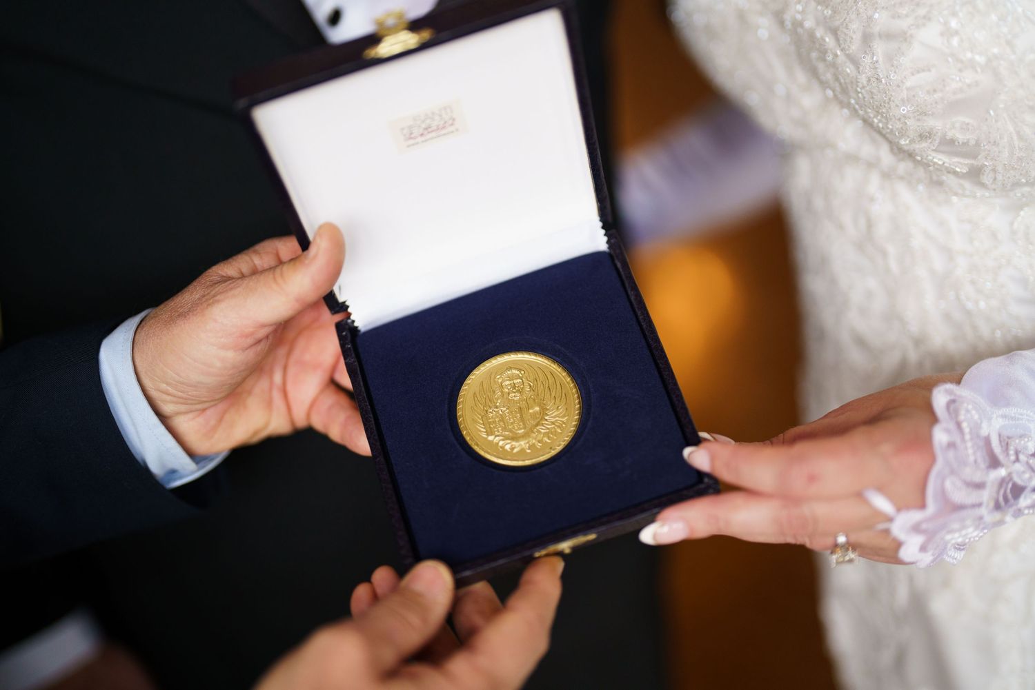 Hands holding an open navy blue presentation box containing a gold medallion or award medal against a blurred background.