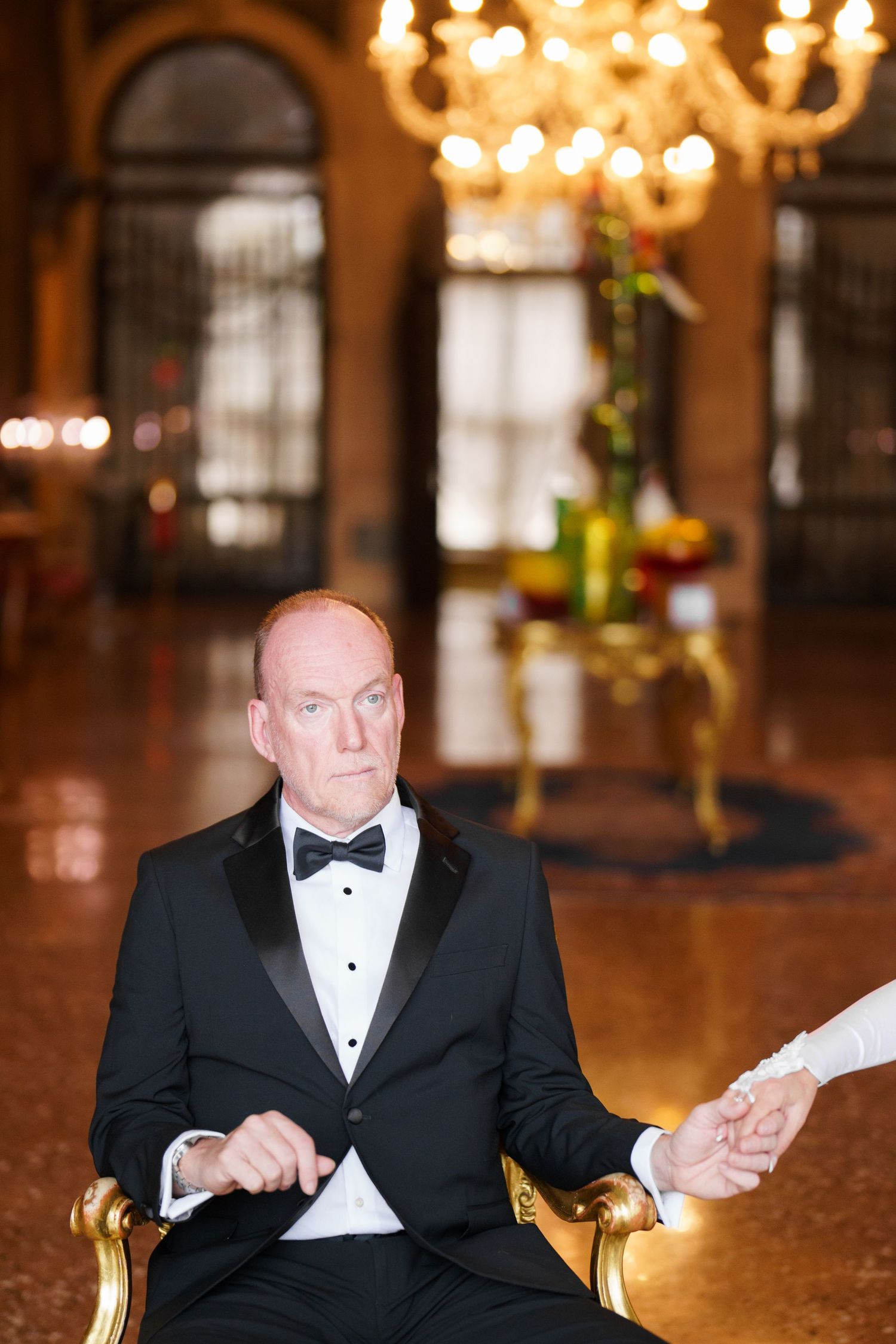 A person in a black tuxedo and bow tie sits at a bar counter under elegant chandelier lighting.