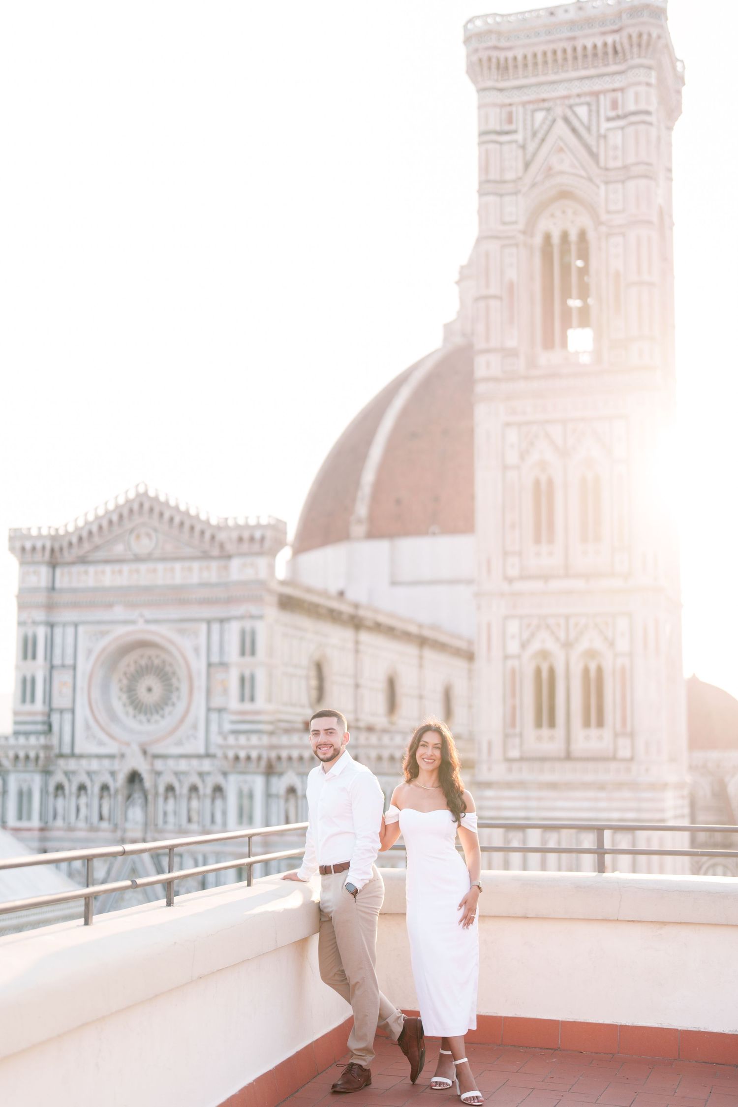 Couple in white outfits standing on rooftop terrace overlooking Florence Cathedral during golden hour in Italy.