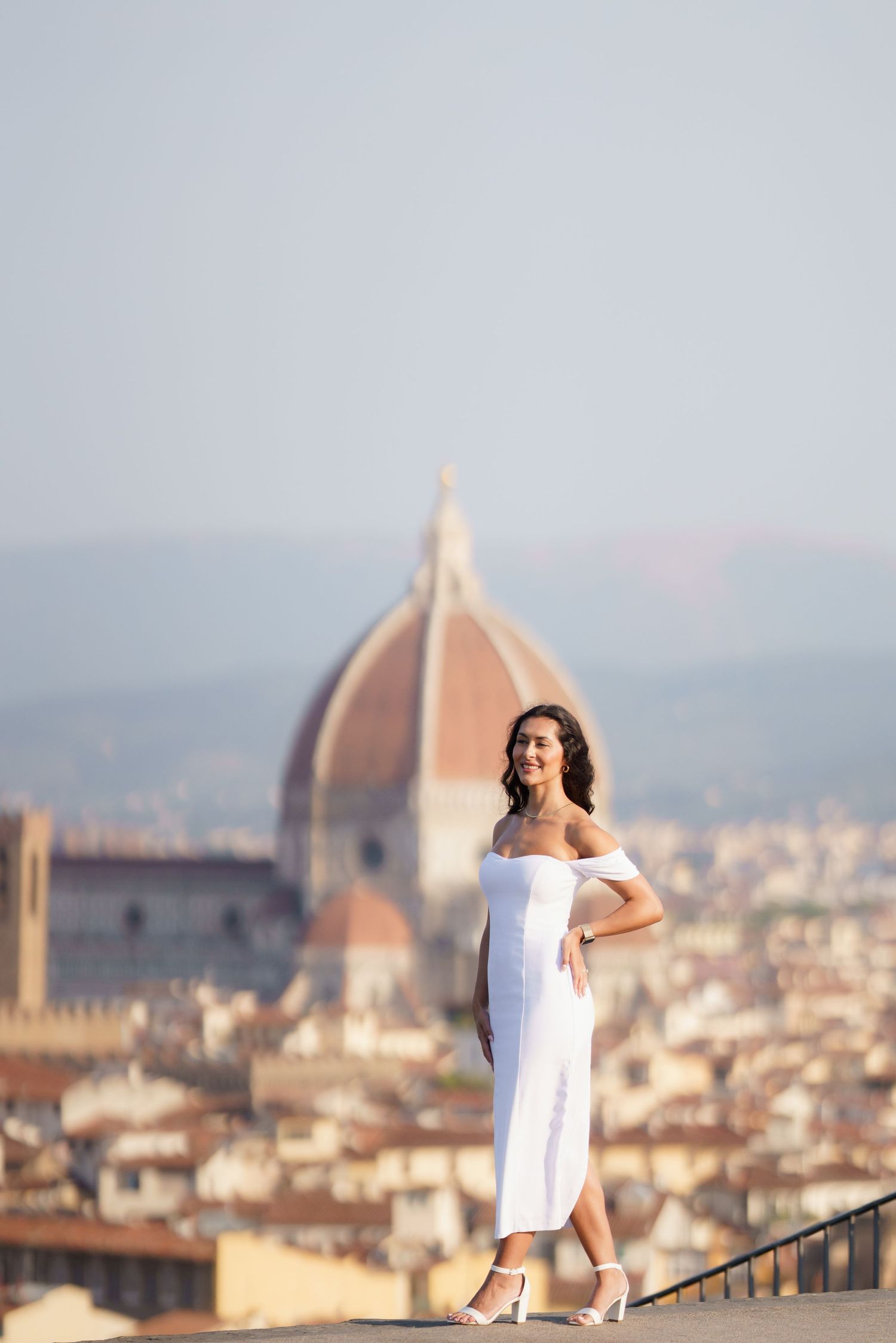 Woman in white dress poses on overlook with Florence's Duomo cathedral dome visible in cityscape background.