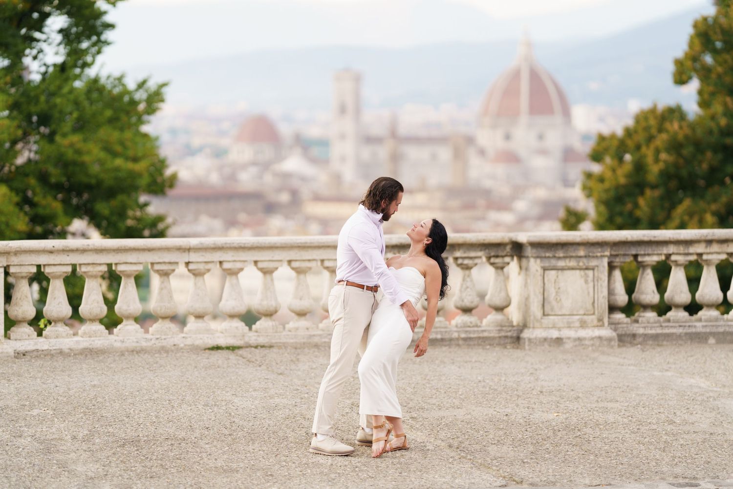 A romantic couple shares a tender moment at a scenic overlook with Florence's Duomo cathedral visible in the distance.