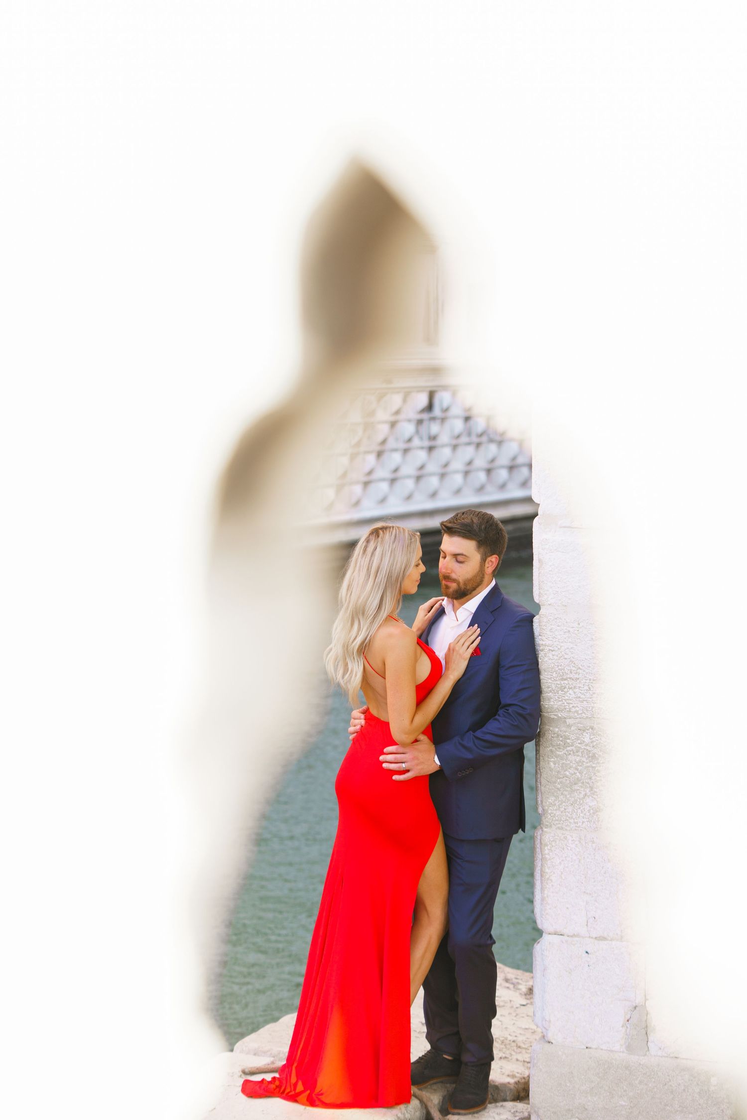 A couple embraces romantically against a white wall near the ocean, with the woman wearing a flowing red dress.