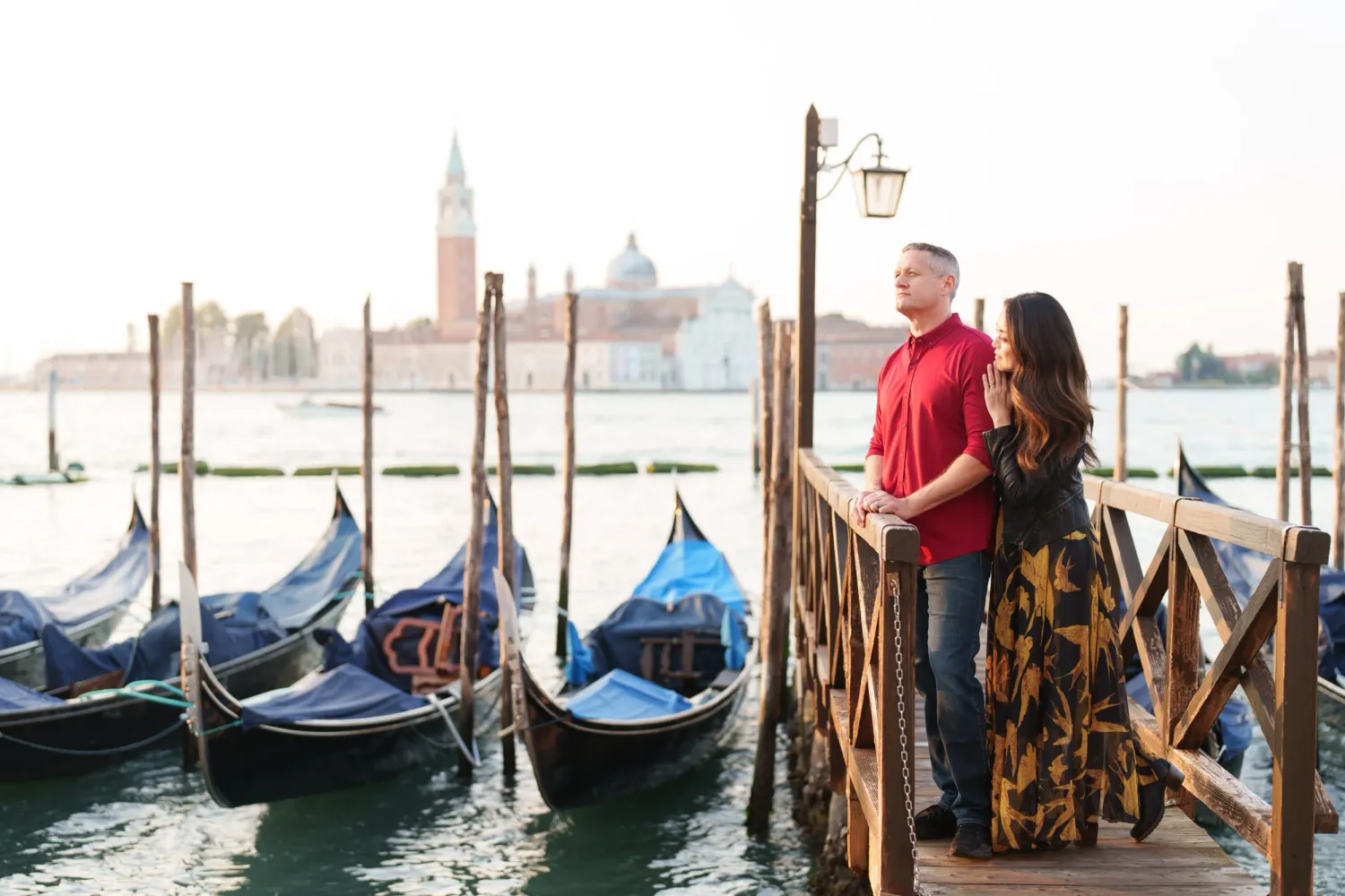 Row of traditional Venetian gondolas moored along wooden posts in Venice lagoon at sunset with city skyline in background.
