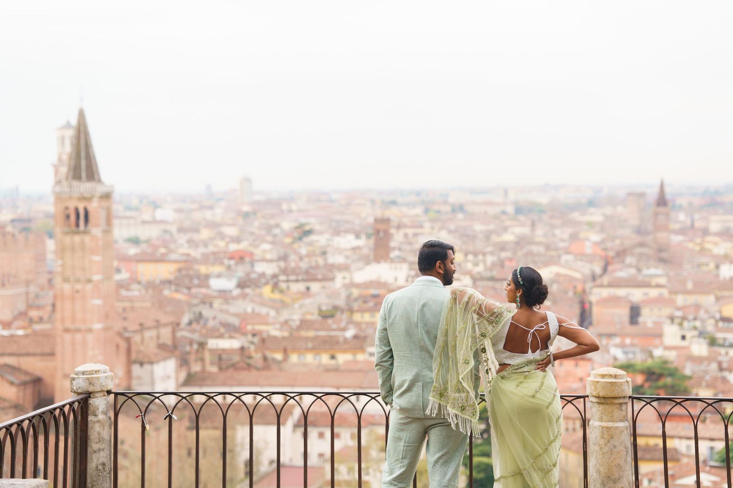 Two people stand at a scenic overlook admiring the panoramic view of Bologna's historic red-roofed cityscape in Italy.