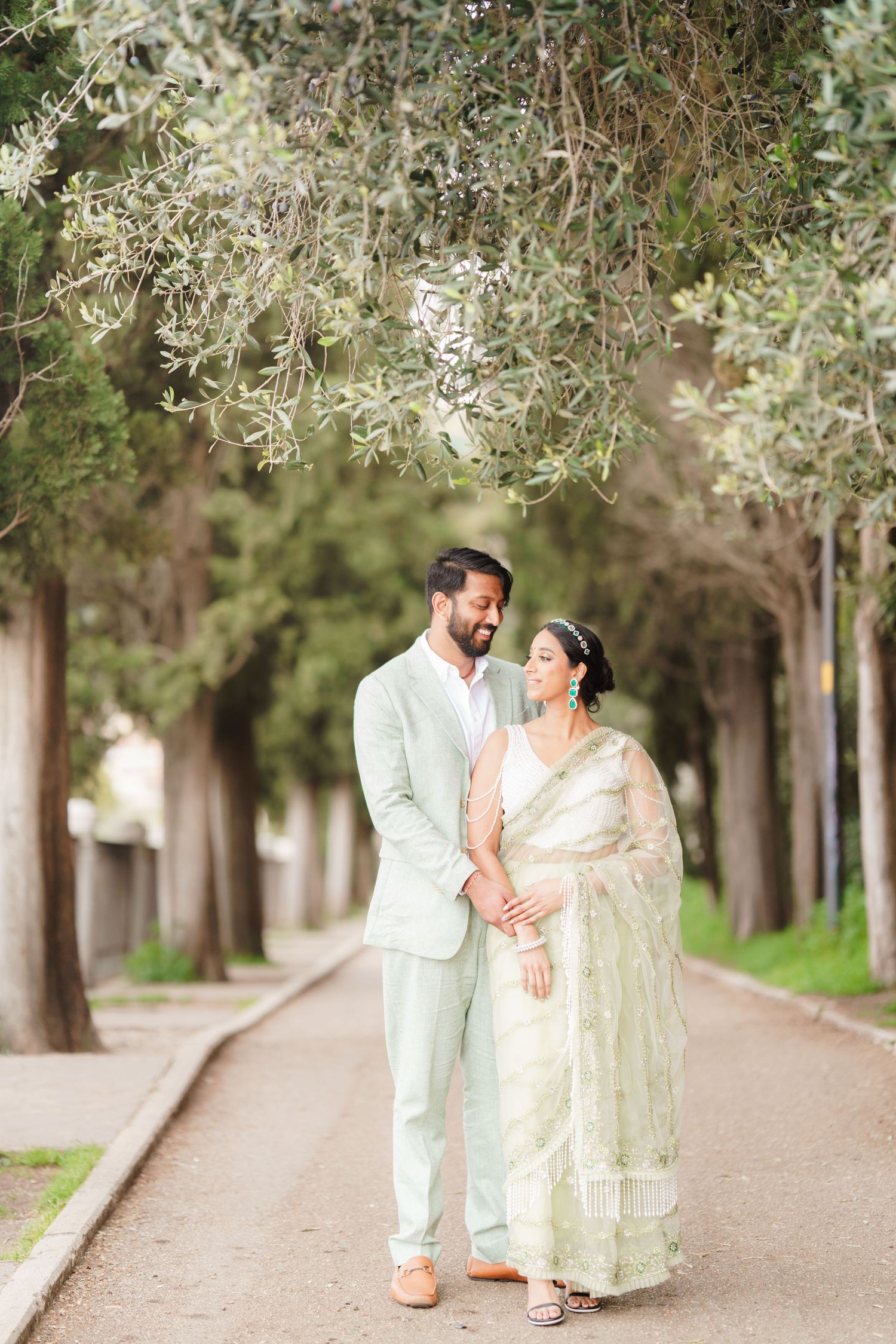 A couple in traditional Indian attire walks together under olive trees on a tree-lined path.