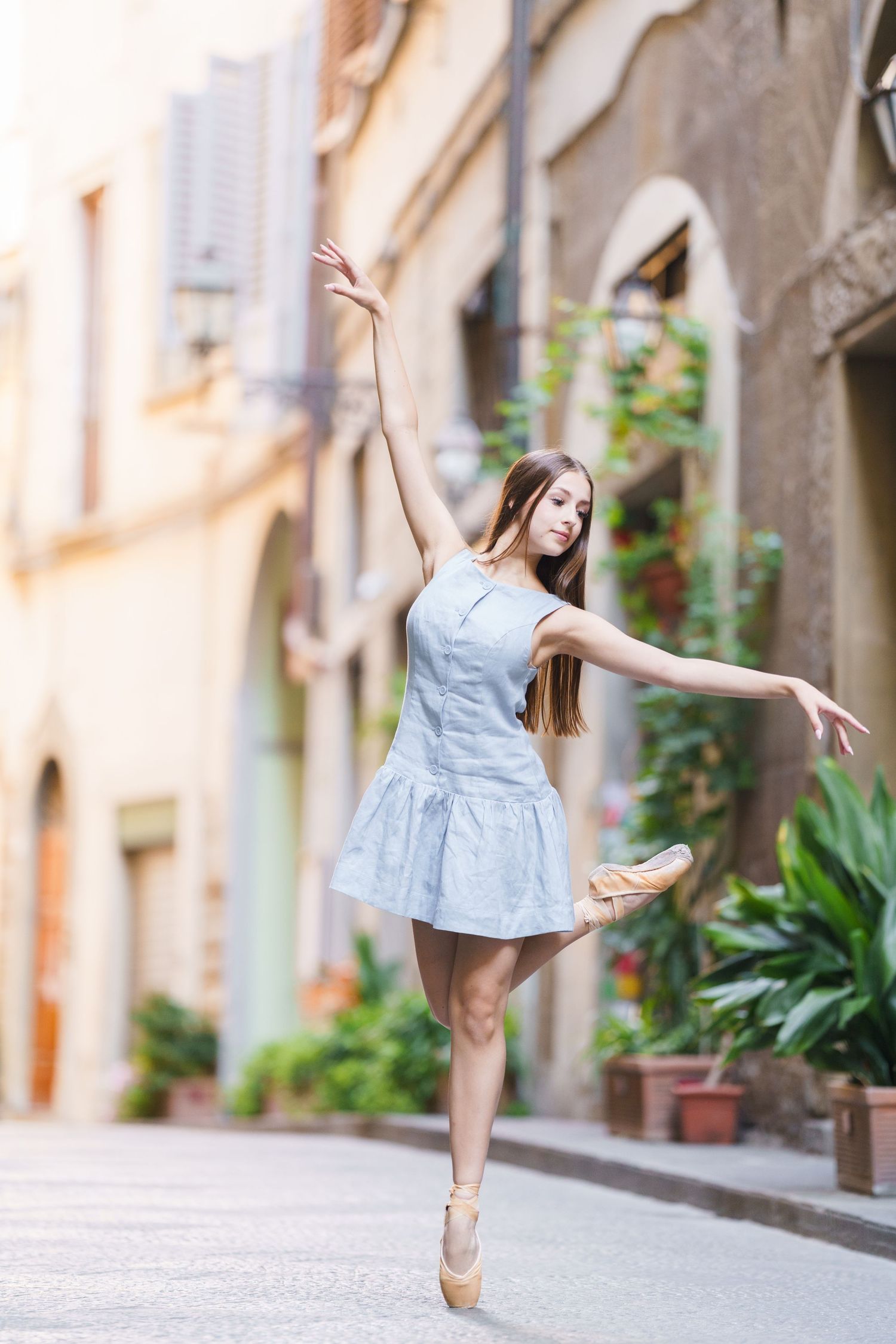A ballet dancer in a light blue dress performs an elegant pose en pointe on a charming European street.