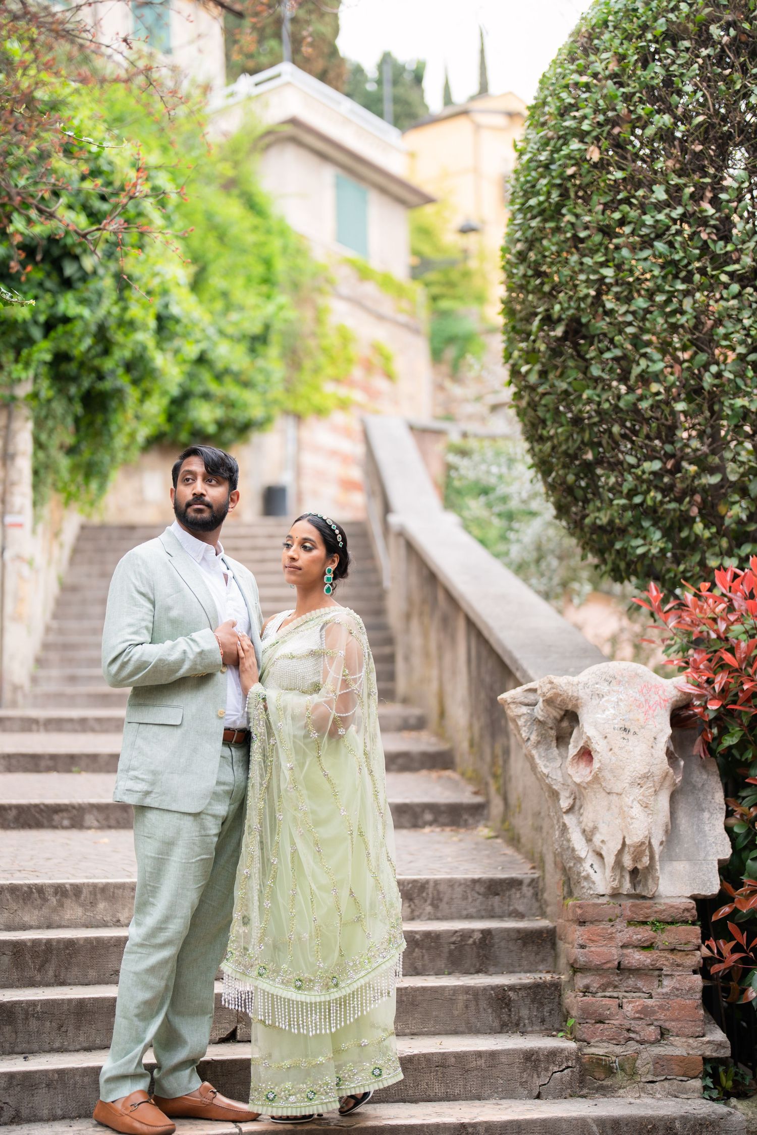 A couple in traditional South Asian attire pose together on stone steps surrounded by lush greenery and flowers.