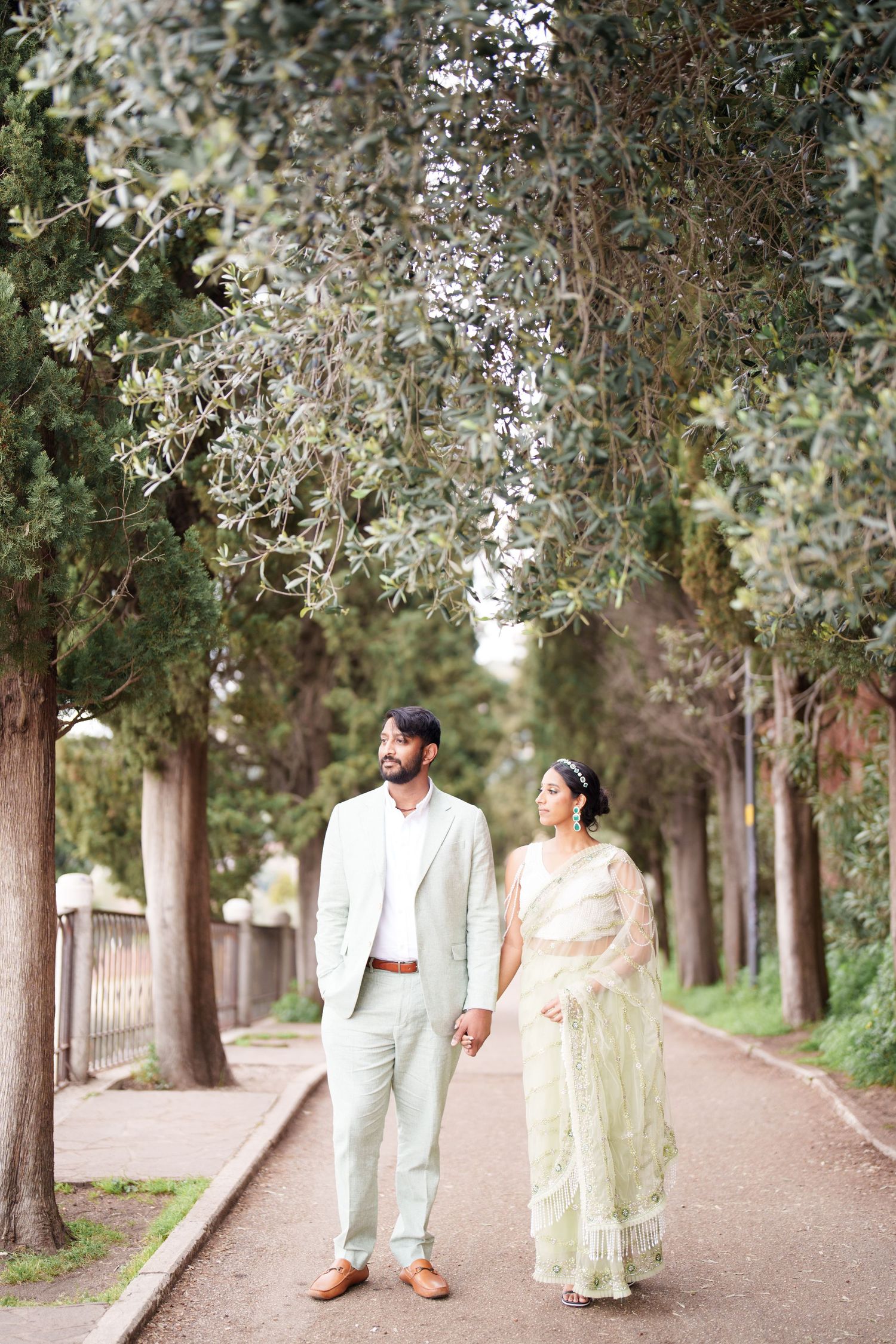 A couple walks hand in hand down a tree-lined path in elegant traditional attire on their wedding day.