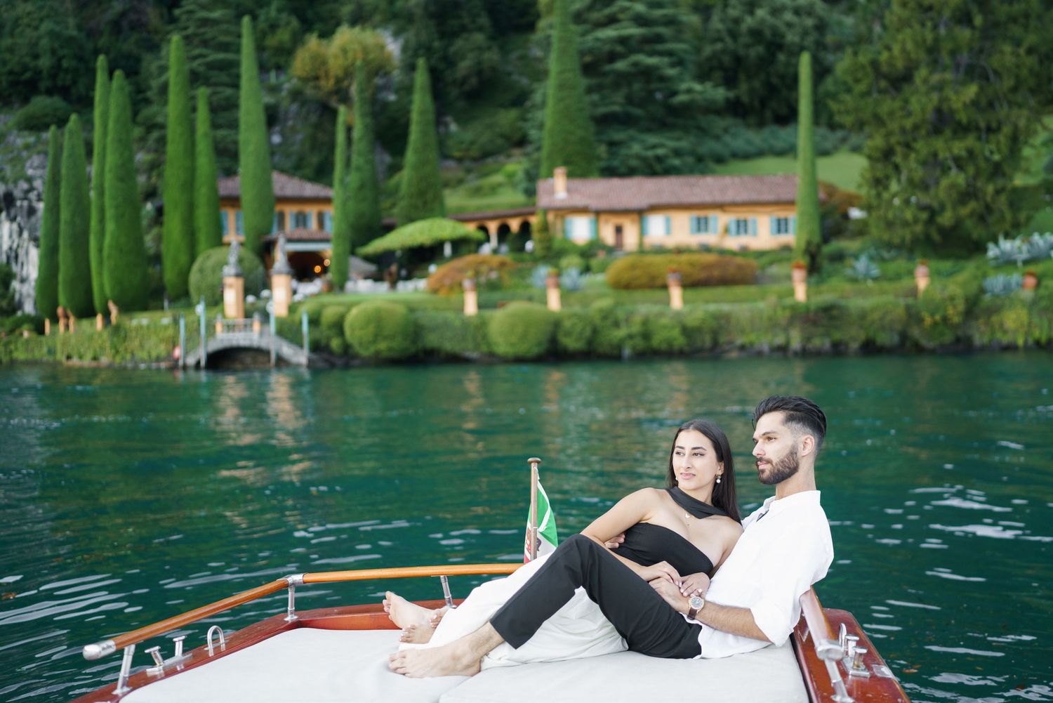 Couple relaxing together on a wooden boat with cypress trees and lakeside villa in the background.