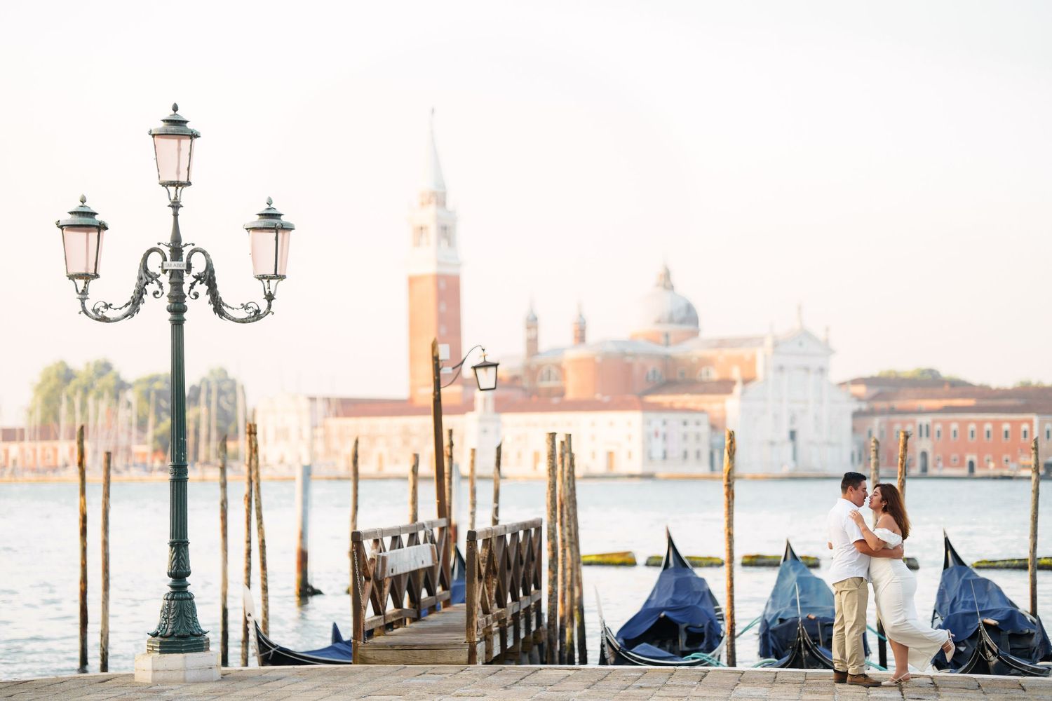 photographer venRomantic couple standing on Venice waterfront with San Giorgio Maggiore church and ornate street lamps in soft sunset light.