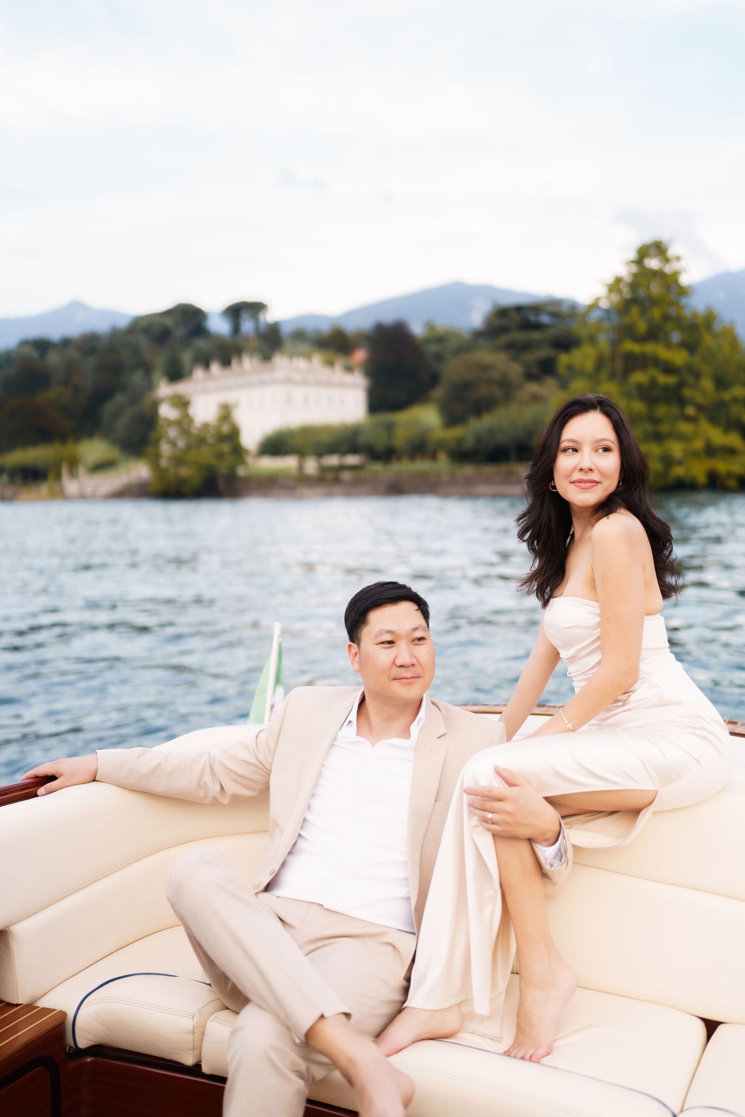 Two people relax on plush cream-colored cushions aboard a luxury boat with mountains and water in the background.