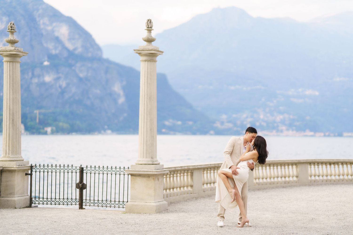 A couple embraces on an elegant lakeside terrace with white stone columns and mountain views at Lake Como, Italy.
