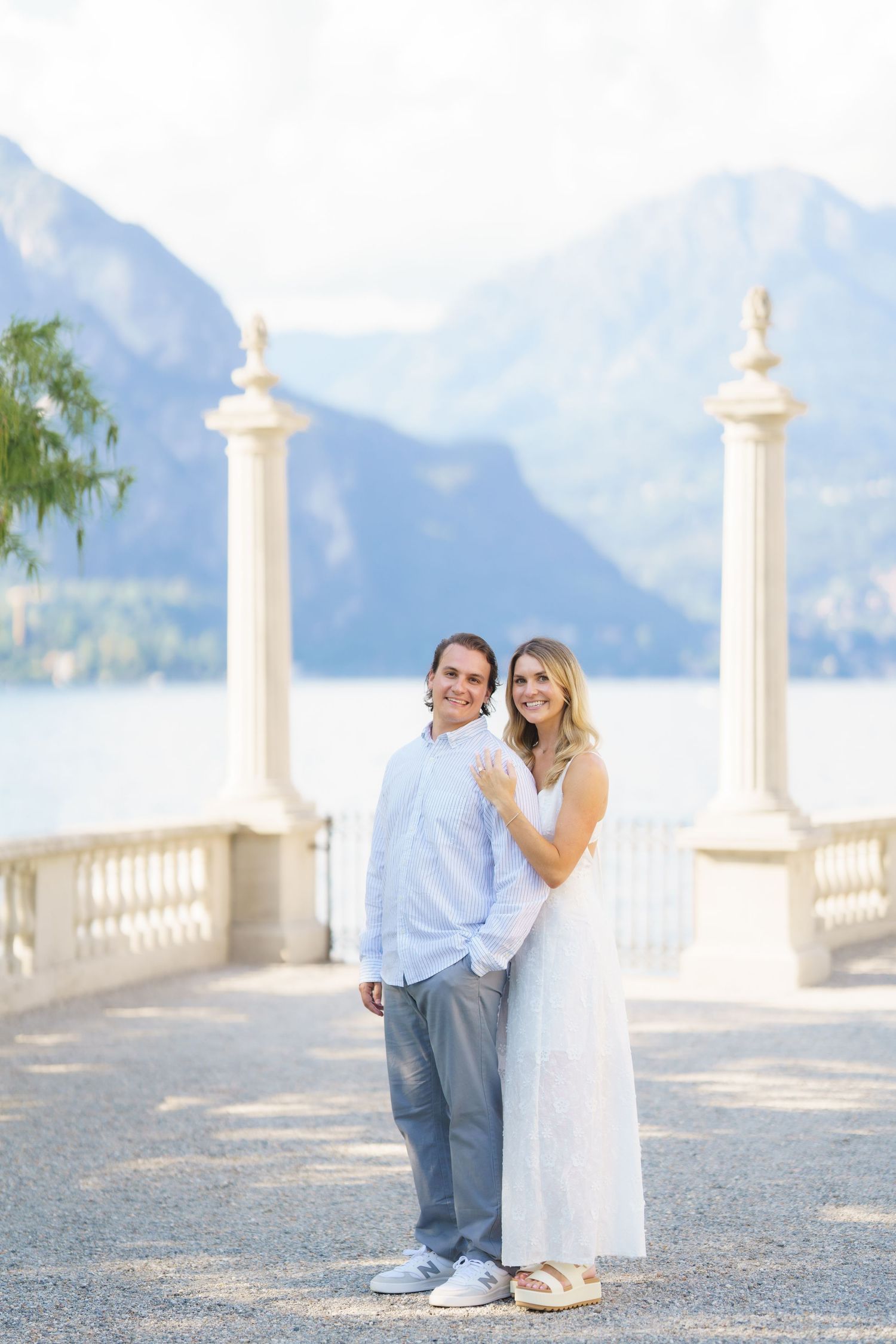 Romantic couple embraces on a scenic terrace overlooking mountains and lake with white stone columns in Italy.