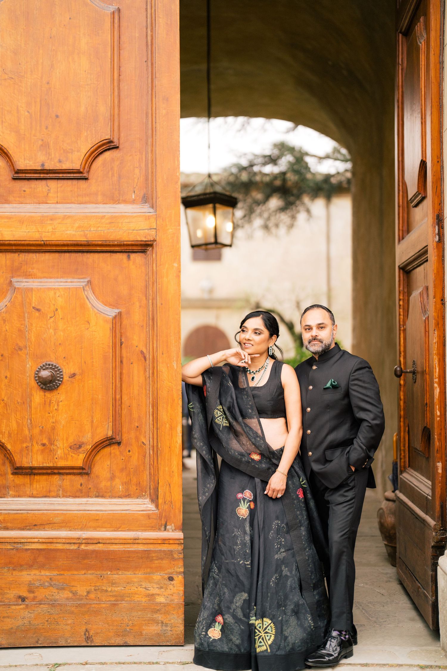 Elegant couple poses in formal black attire under an ornate archway with wooden doors.