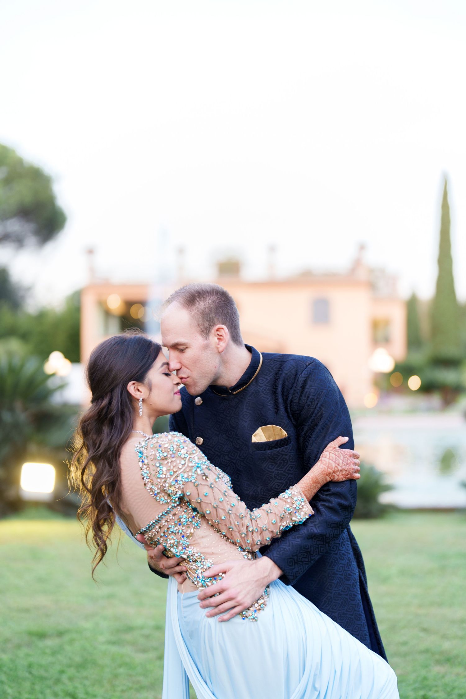 Couple embracing in an elegant outdoor setting with a Mediterranean villa in the background.
