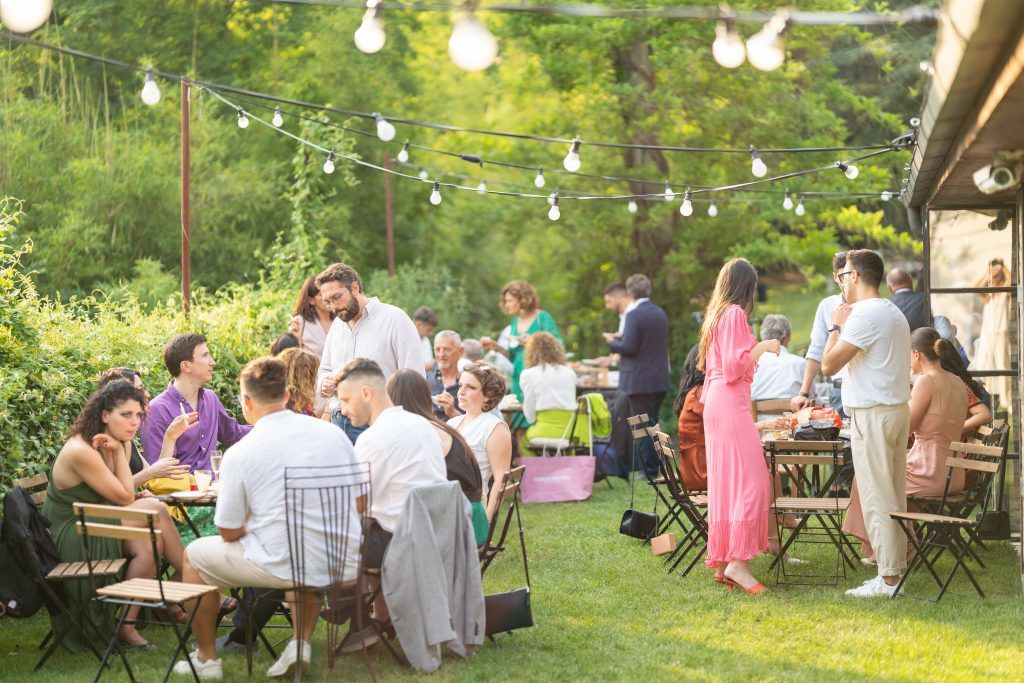 String lights illuminate an outdoor reception area with picnic tables and mingling guests.