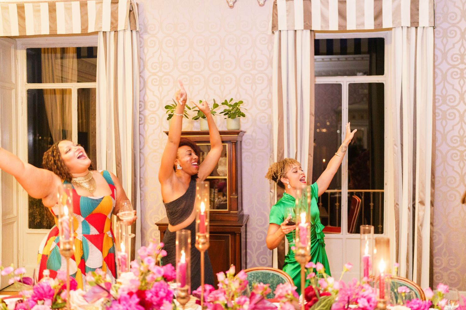 Three women dancing joyfully at an elegantly decorated dinner table with colorful flowers and candles.