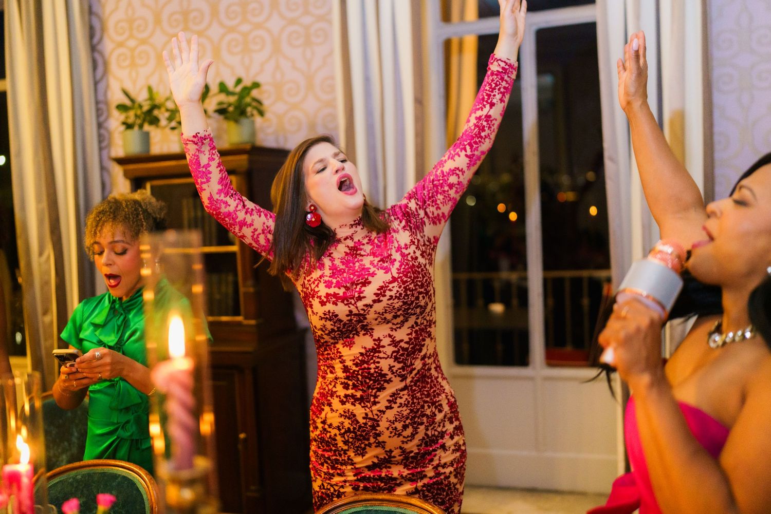 Women in sequin dresses celebrate at an elegant party, raising hands joyfully near candlelit tables.